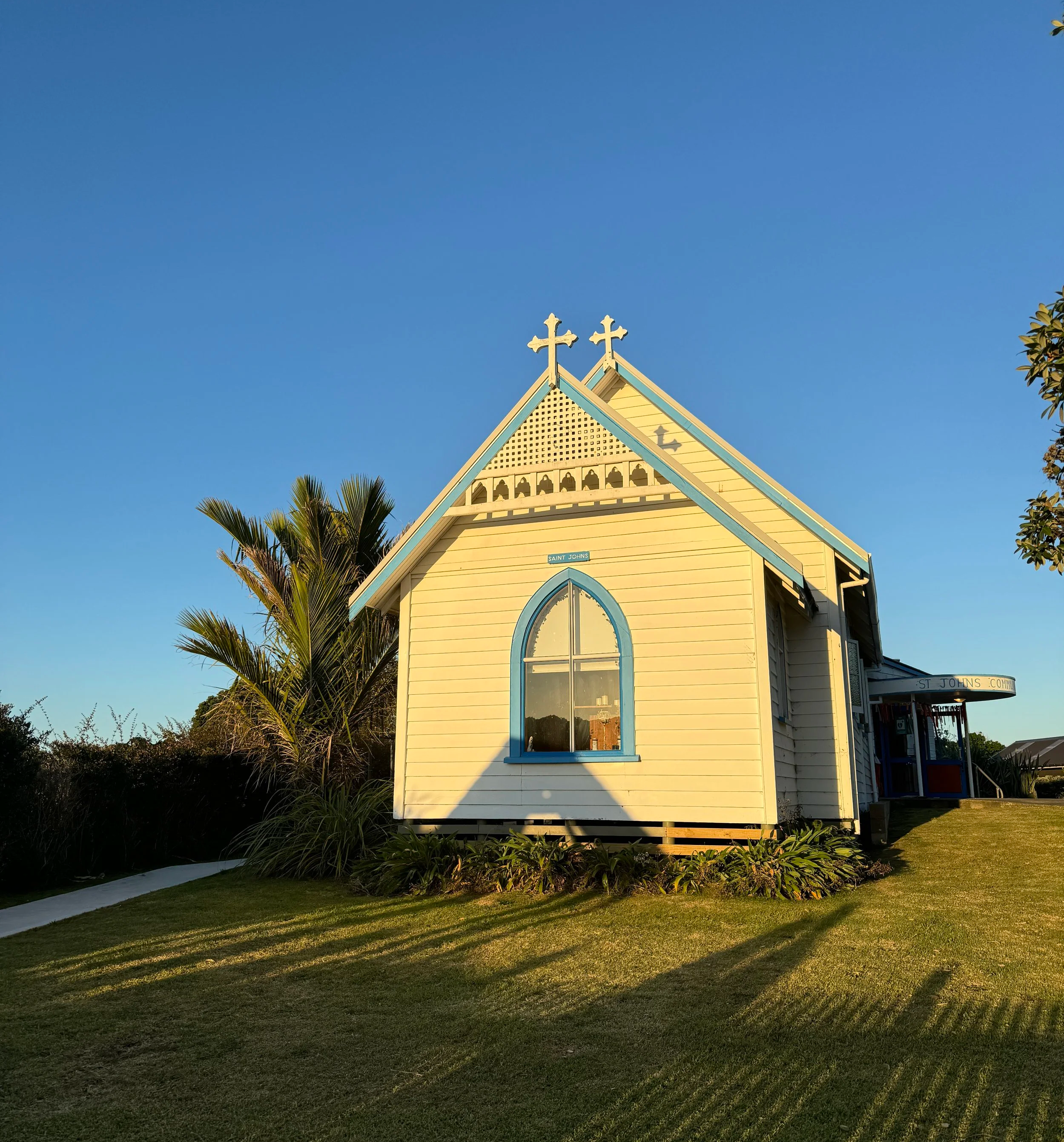 A small yellow church with blue accents and two crosses on top, surrounded by palm trees and shrubs under a clear blue sky.