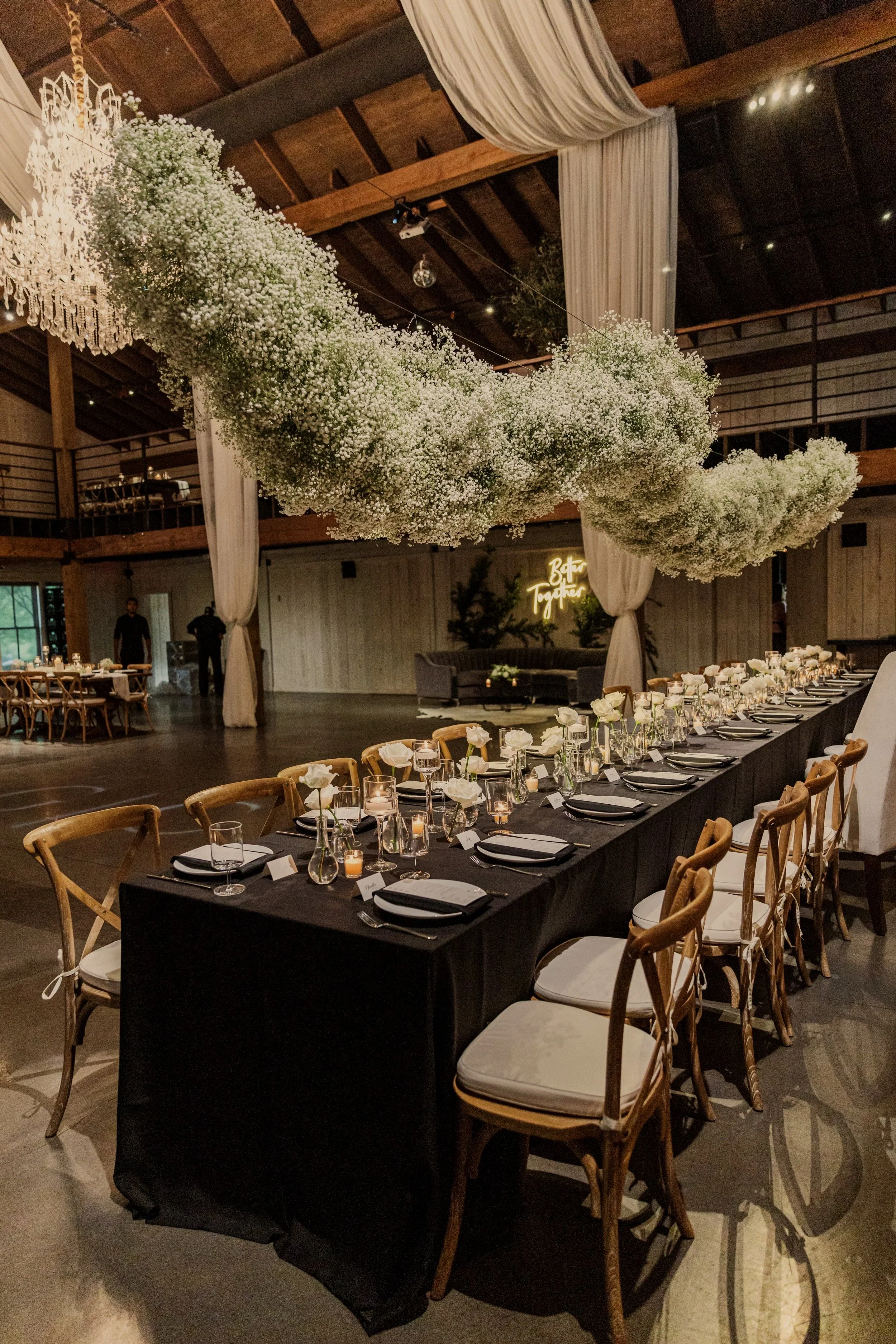A wedding reception setup with a long table covered with a black tablecloth, set with plates, glasses, and small candles. The chairs are wooden with white cushions. Above the table, there is a large cloud-like arrangement of white baby's breath flowers, and a wooden ceiling with draped white fabric. A neon sign in the background reads