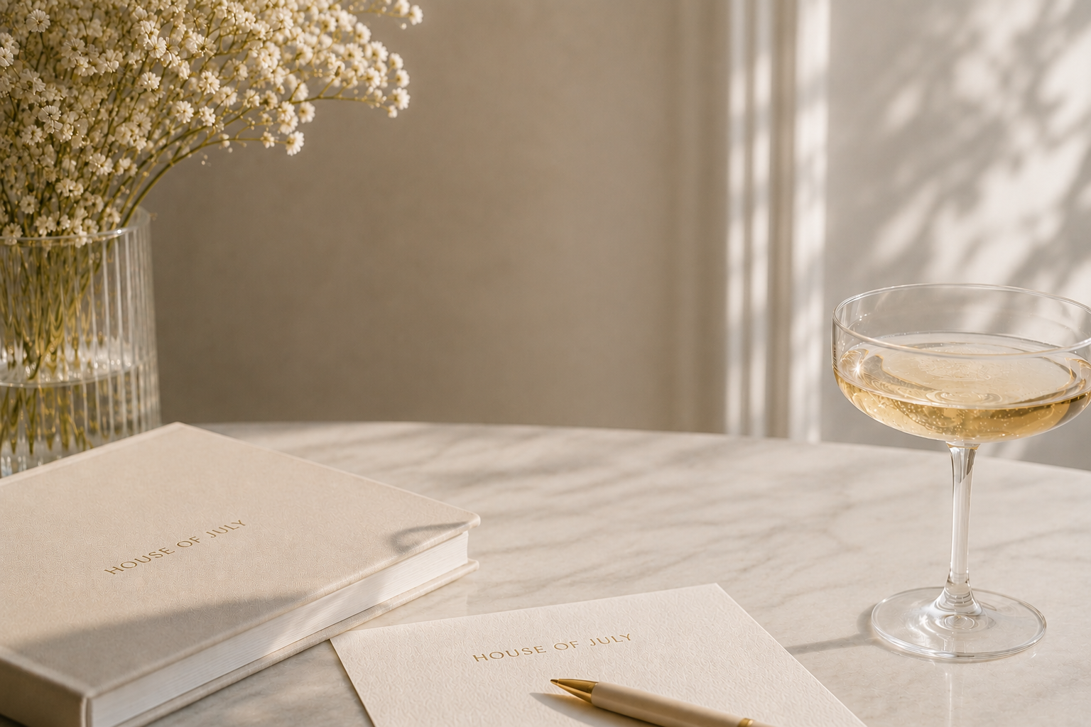 A table with a white cover, a glass of white wine or champagne, a closed notebook titled 'House of July', a matching pen, and a white vase with white daisies. Shadows of the daisies are cast on the wall.