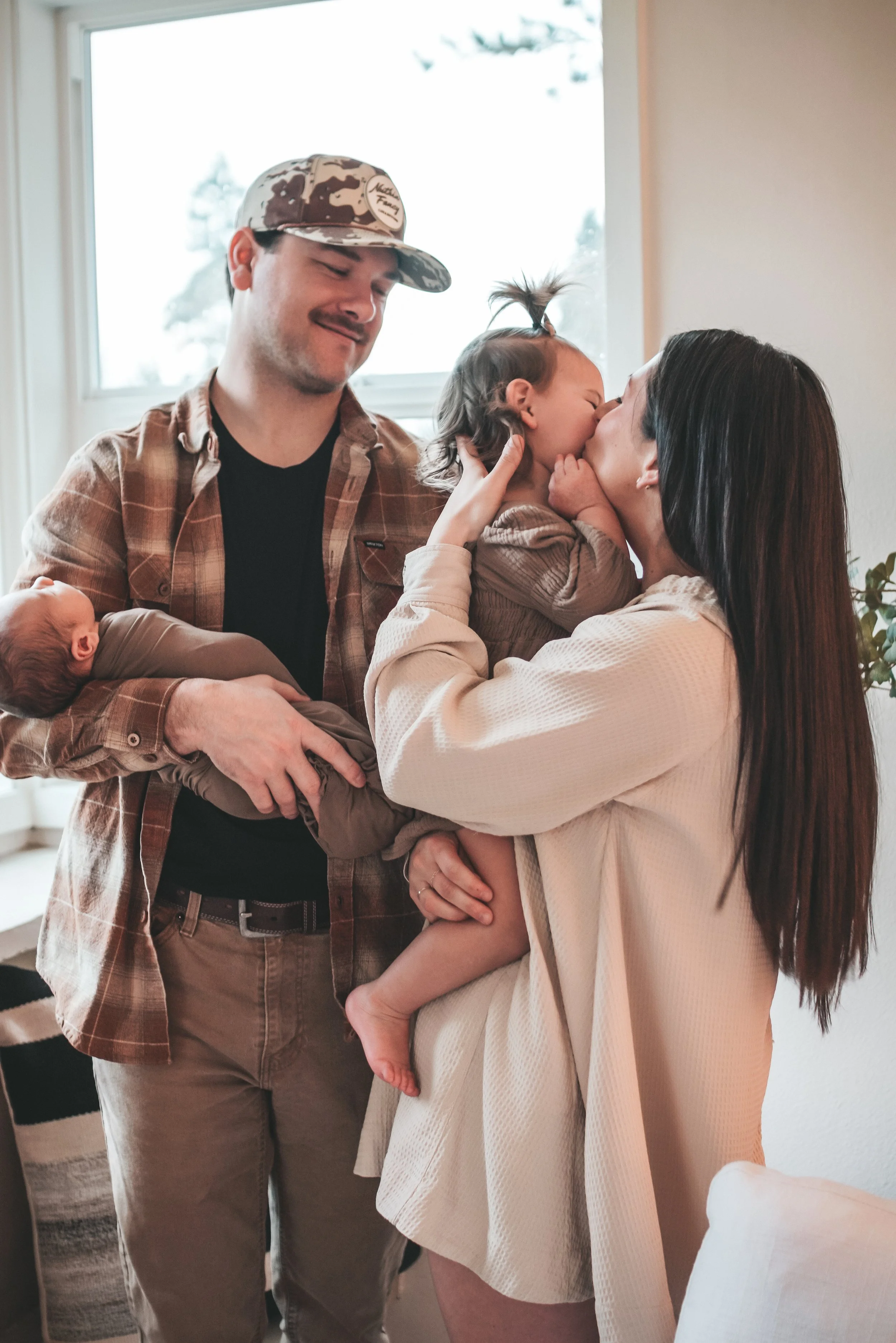A family gathers inside a bright, modern home celebrating the arrival of a new baby. The mother is holding an older child, who is kissing the baby's face, and the father is holding a newborn wrapped in a blanket, looking delighted.