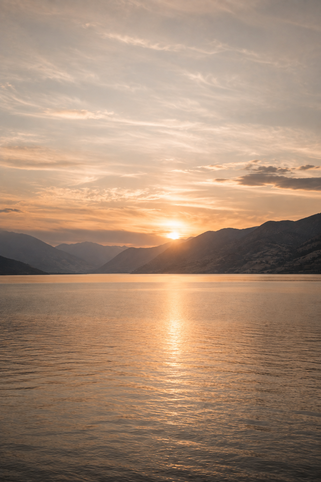 Sunset over a calm lake surrounded by mountains with a partly cloudy sky.