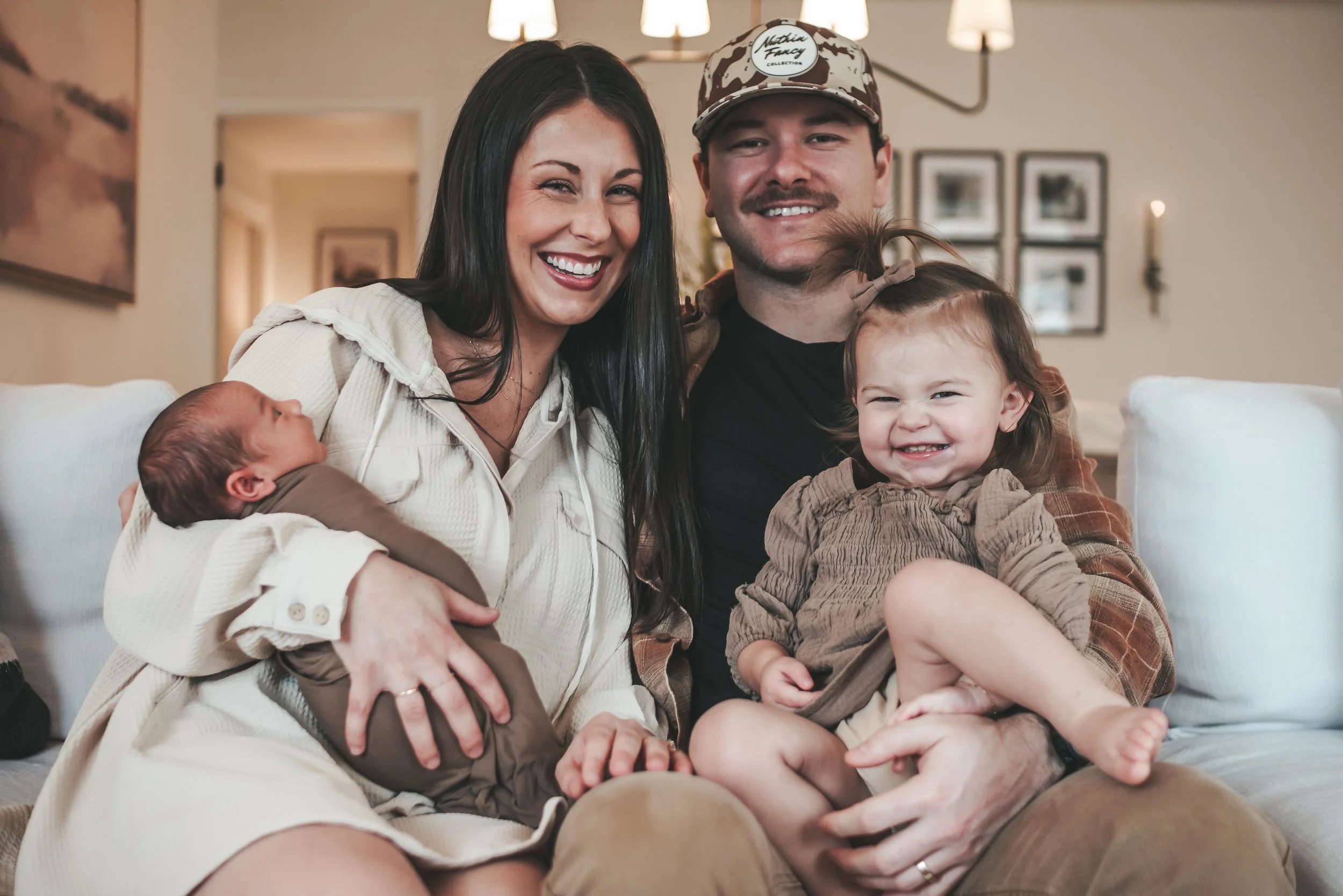 A family of four sitting on a couch in a cozy living room. The mother, with long black hair, is holding a newborn baby wrapped in a brown blanket. The father, wearing a camouflage cap and black shirt, is smiling at the camera. A young girl with curly hair and a plaid dress is sitting in the father’s lap, smiling happily. The background features framed pictures and warm lighting.