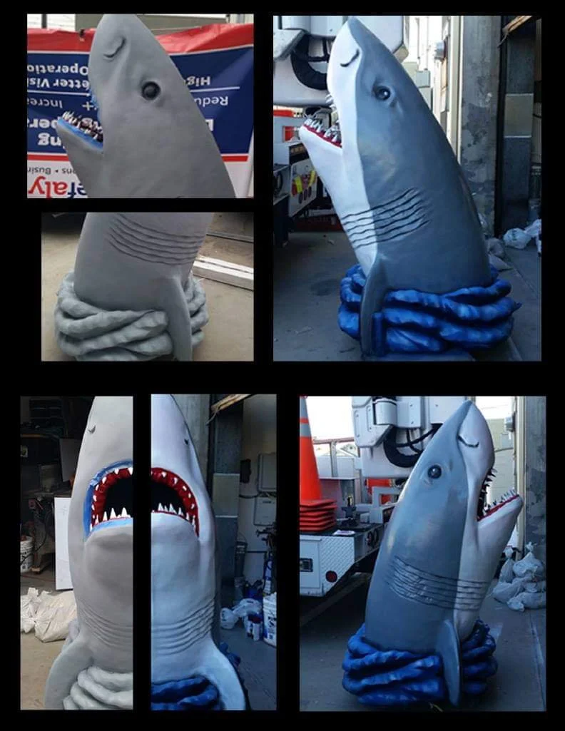 A collage of four images showing a large shark costume. The costume includes a shark head with an open mouth and sharp teeth, a body with detailed gills, and a circular base that looks like blue water or waves. The costume is displayed both indoors and outdoors, with some images showing the shark standing upright and one with the mouth wide open.