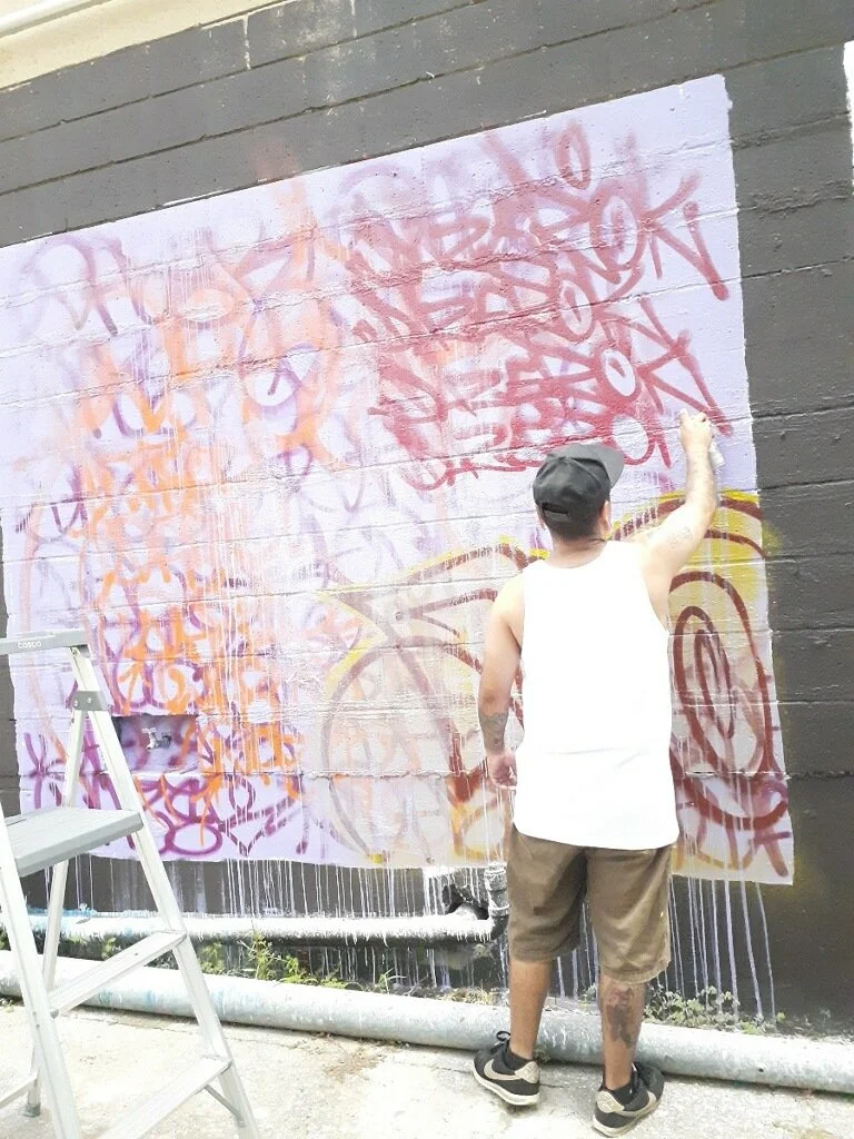 Person wearing a white sleeveless shirt and brown shorts spray-painting graffiti on a large wall with a ladder nearby.