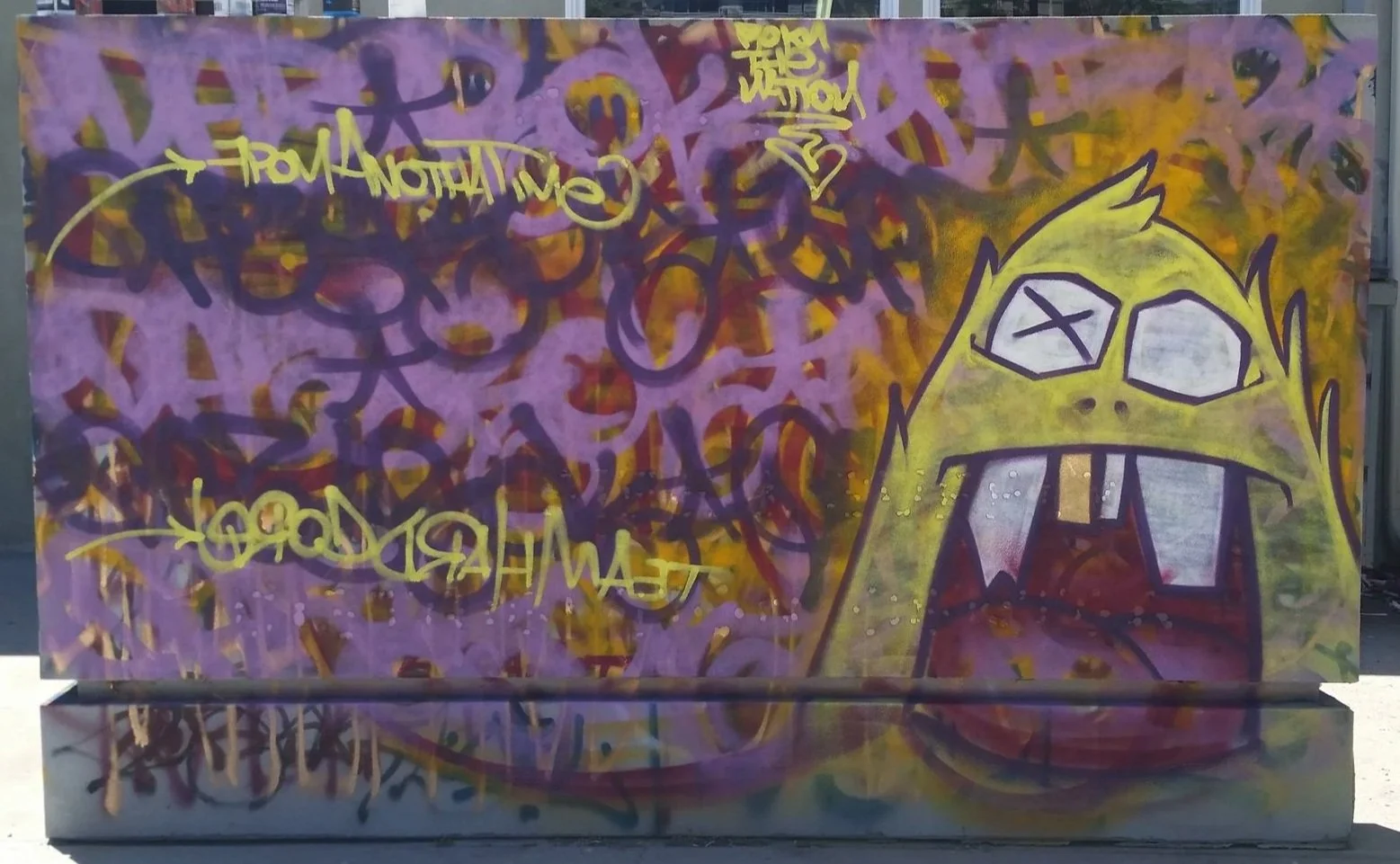 Graffiti art featuring a cartoonish yellow character with glasses, large furrowed eyebrows, and mouths with large white teeth, against a background of swirling purple, red, and yellow graffiti tags and phrases.
