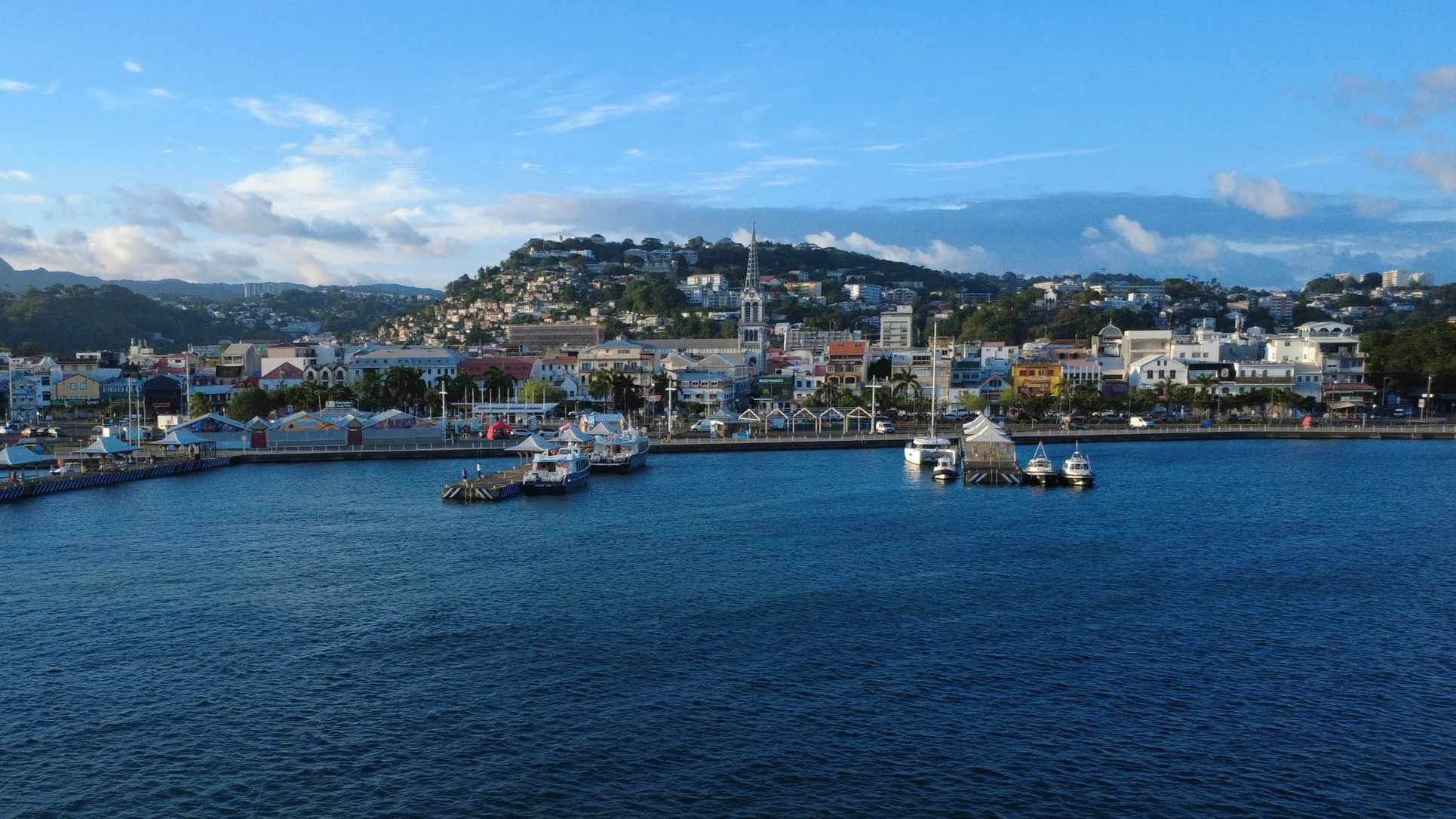 Une vue d'une ville portuaire avec des bateaux amarrés dans le port, des bâtiments colorés et une colline en arrière-plan avec des maisons, sous un ciel partiellement nuageux.