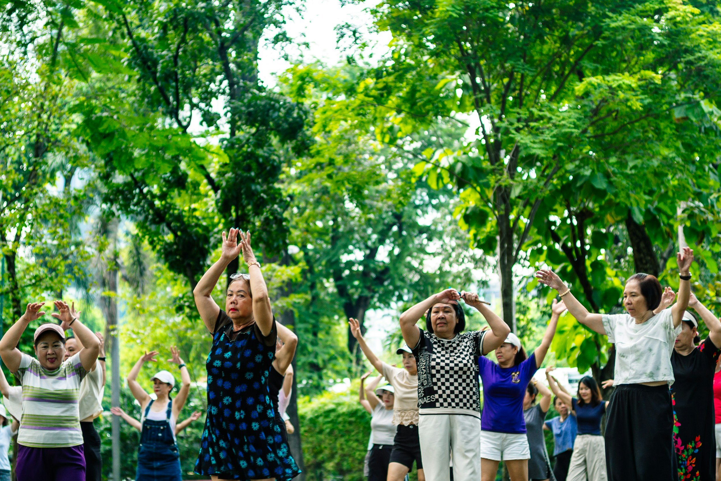 Group of women participating in outdoor exercise class, raising arms, surrounded by green trees.