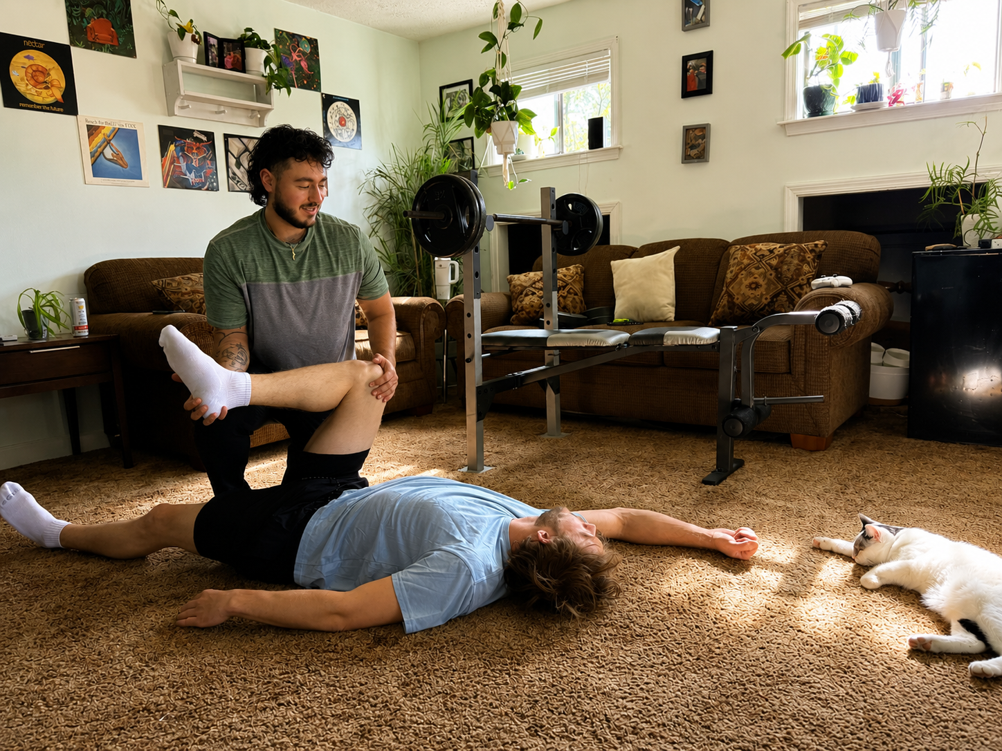 A man lying on the floor with another man kneeling and stretching his leg while holding it. A white and black cat is lying nearby, and the scene is set in a living room with indoor plants, artwork on the walls, a brown sofa with pillows, and a workout bench.