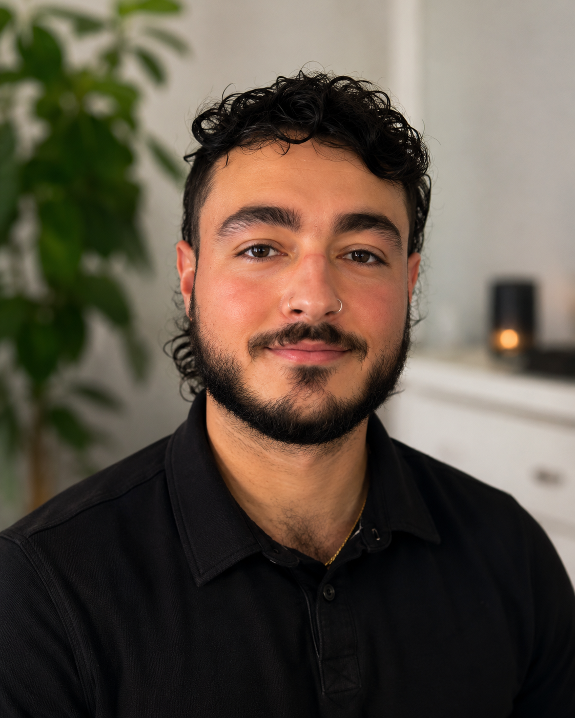 Close-up of a young man with curly black hair, a beard, wearing a black collared shirt, smiling slightly, with a plant and a lit candle on a dresser in the background.
