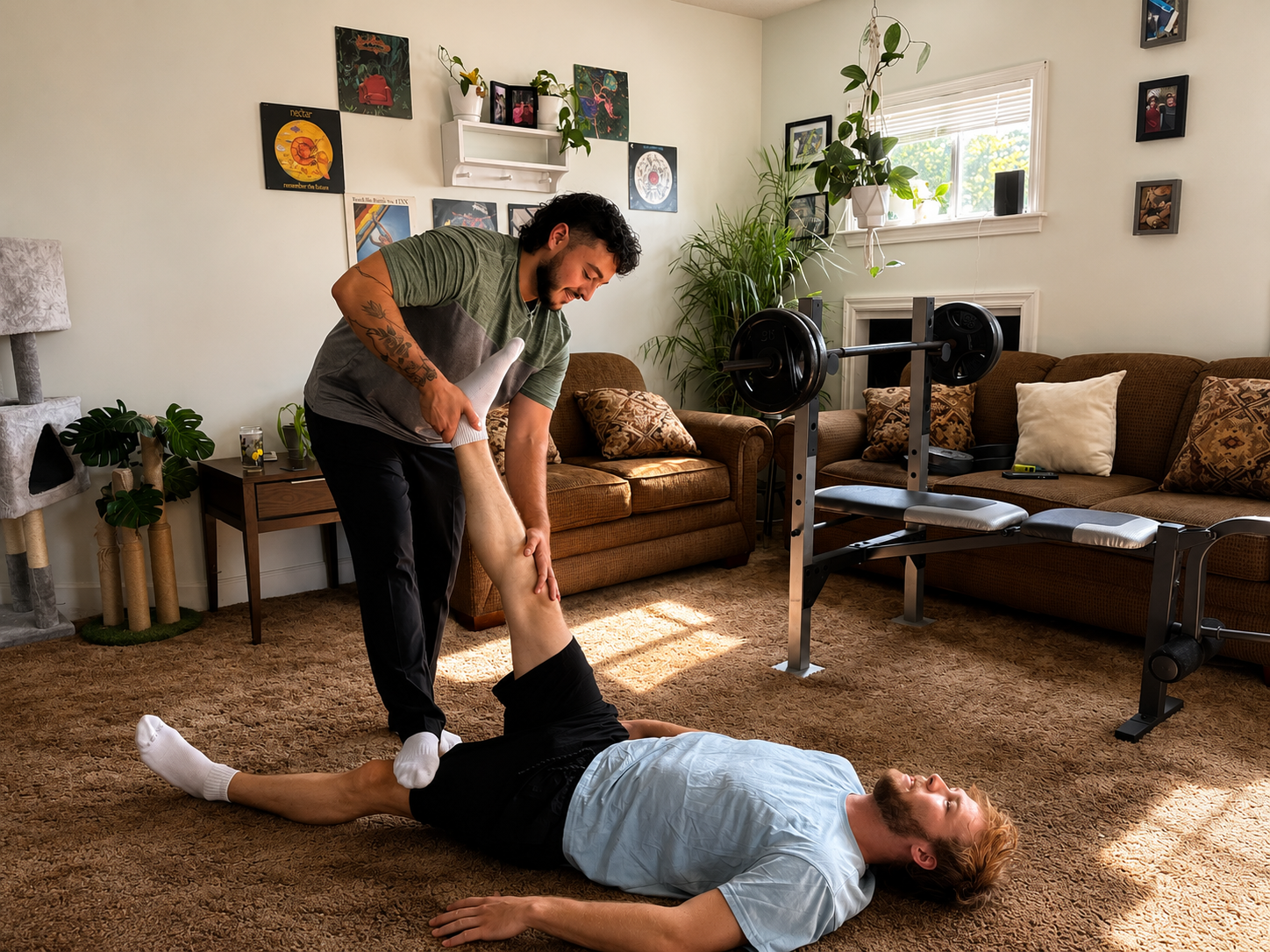 A man lying on the carpet in a living room while another man helps stretch his leg. The room has a brown carpet, a brown couch with patterned pillows, a window with sunlight, and wall art. Fitness equipment and potted plants are also visible.
