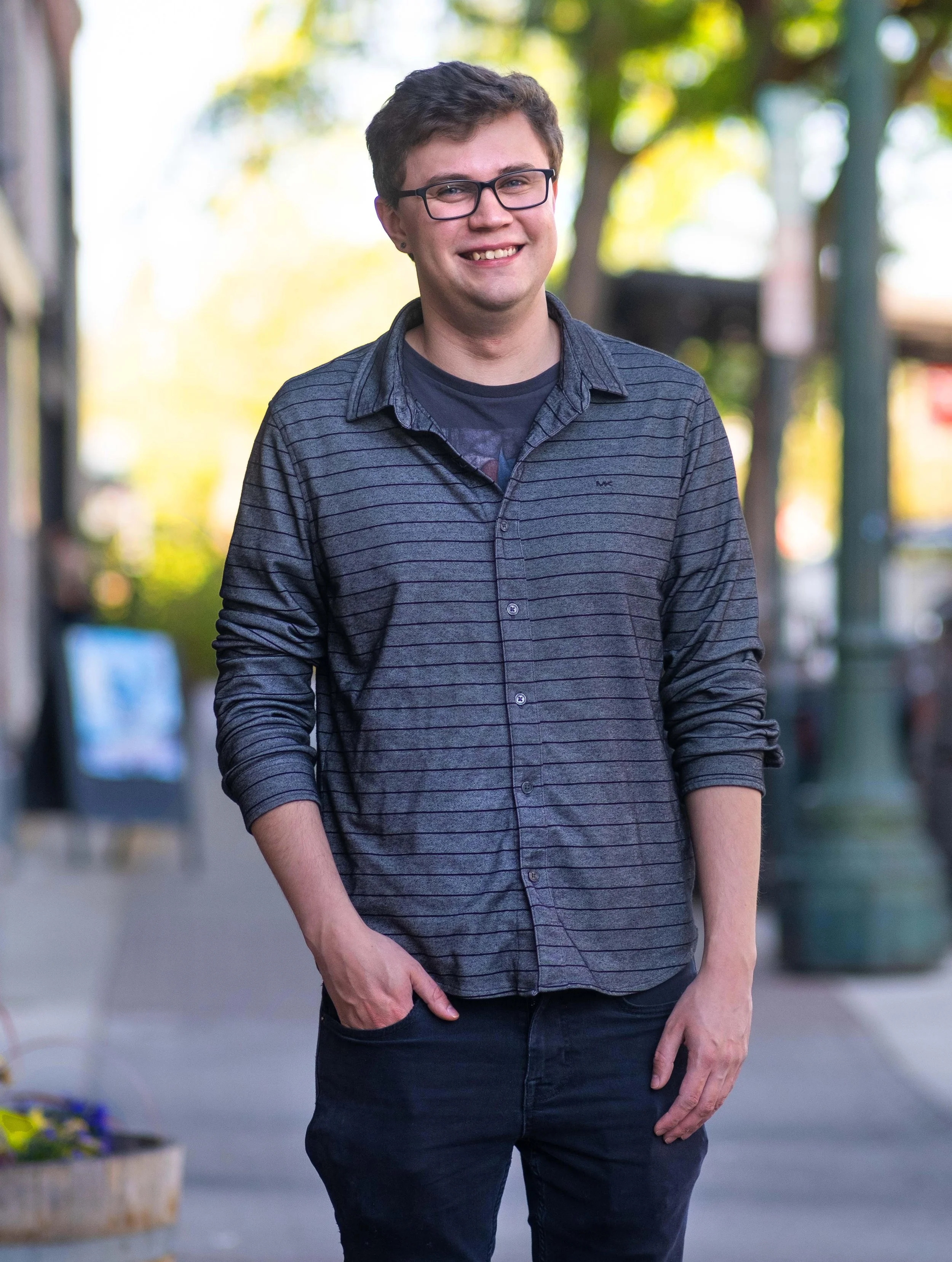 Bronson Moreno smiling while walking outdoors on a street lined with trees with green and yellow leaves, wearing a dark striped shirt and black pants.