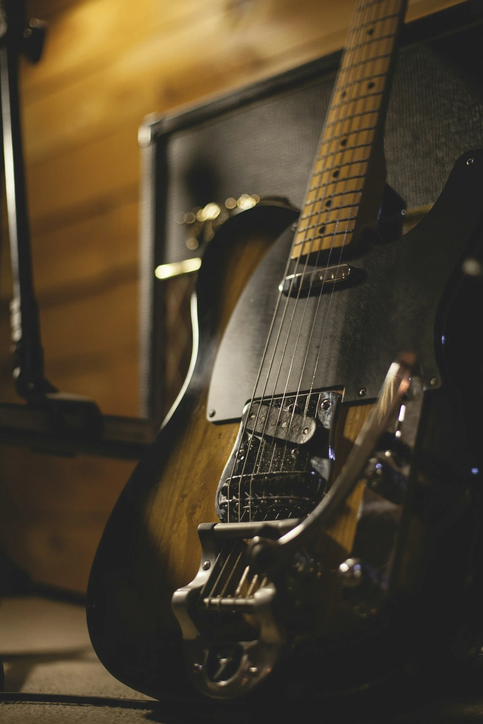 Close-up of an electric guitar leaning on a black amplifier, with a focus on the guitar's body, fretboard, and strings. The background shows a wooden floor and a blurred wall.