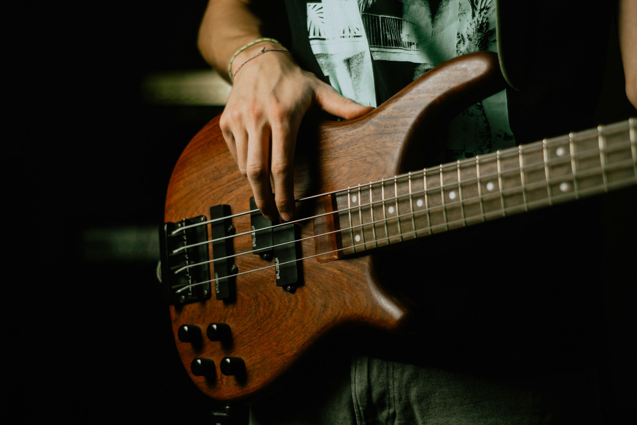 Close-up of a person playing a wooden electric bass guitar, with focus on their hand on the fretboard and the body of the guitar.
