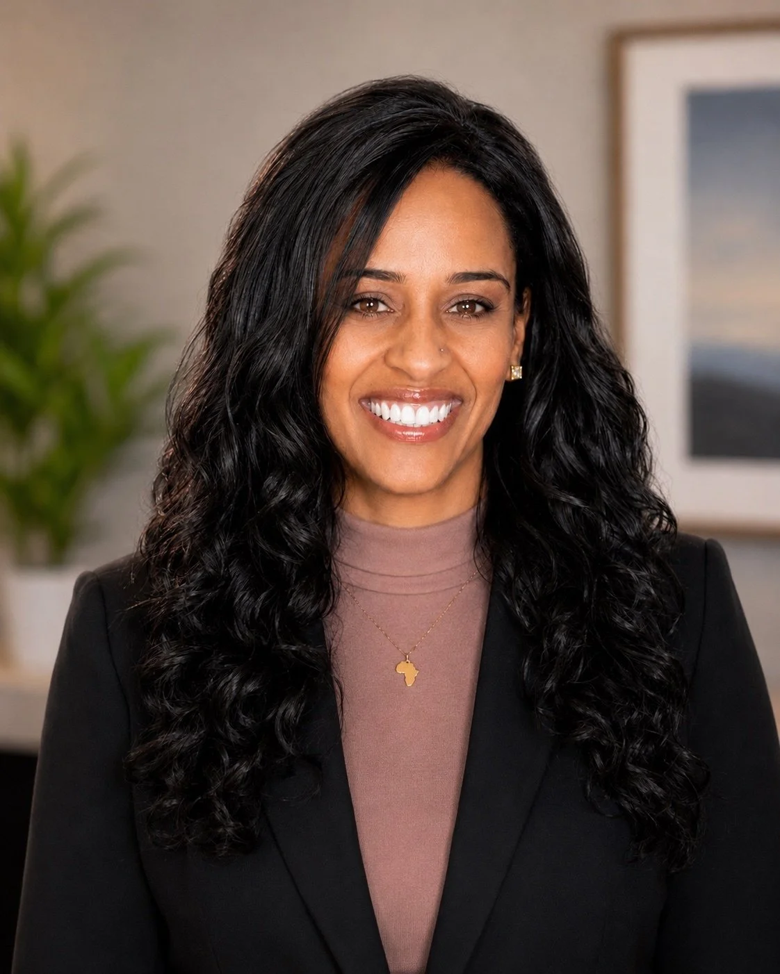 A smiling woman with long, curly black hair, wearing a black blazer and a beige top, standing in an office setting with a plant and framed picture in the background.