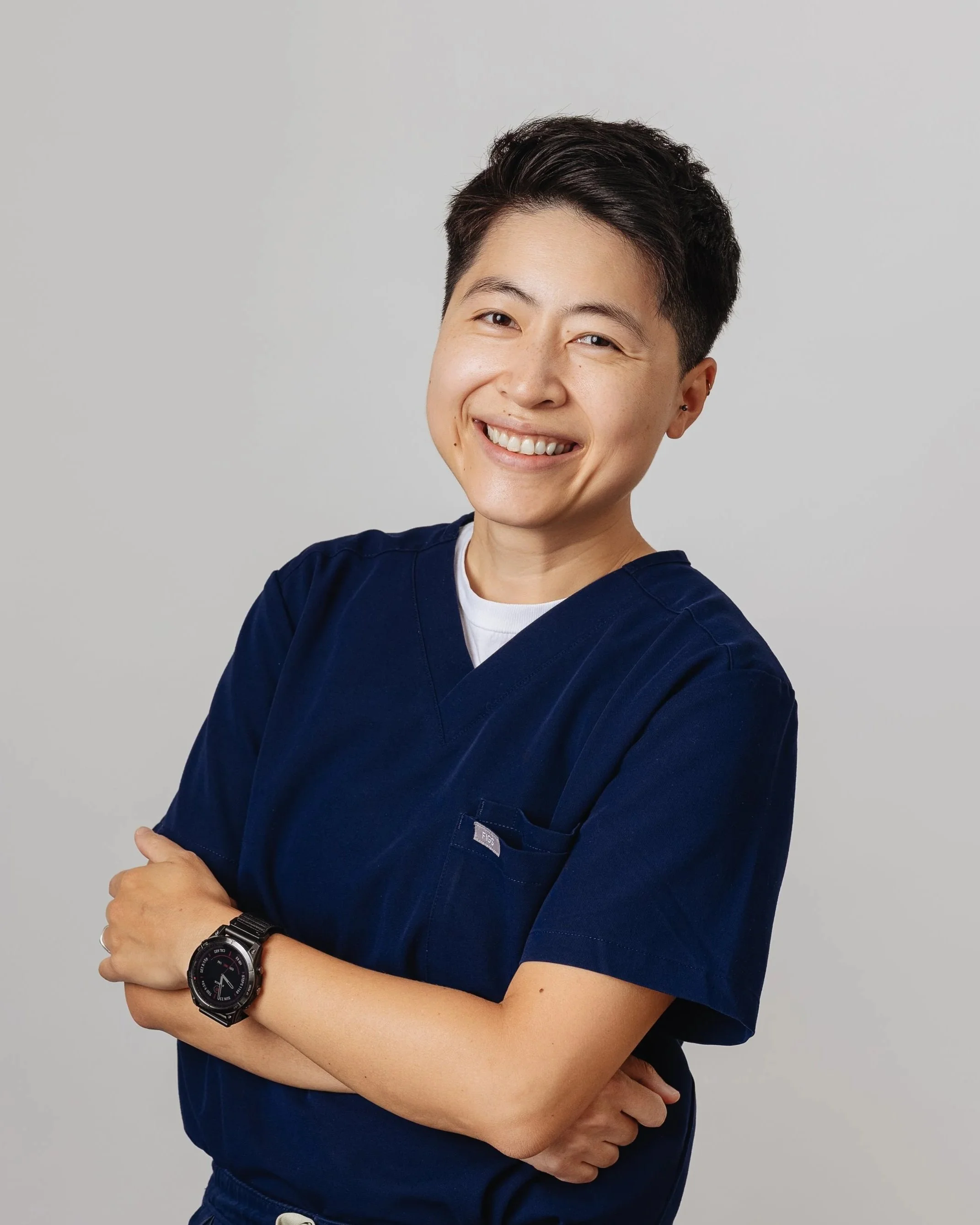 A person wearing a dark blue medical scrub top and a black wristwatch, crossing their arms and smiling at the camera, with a plain gray background.