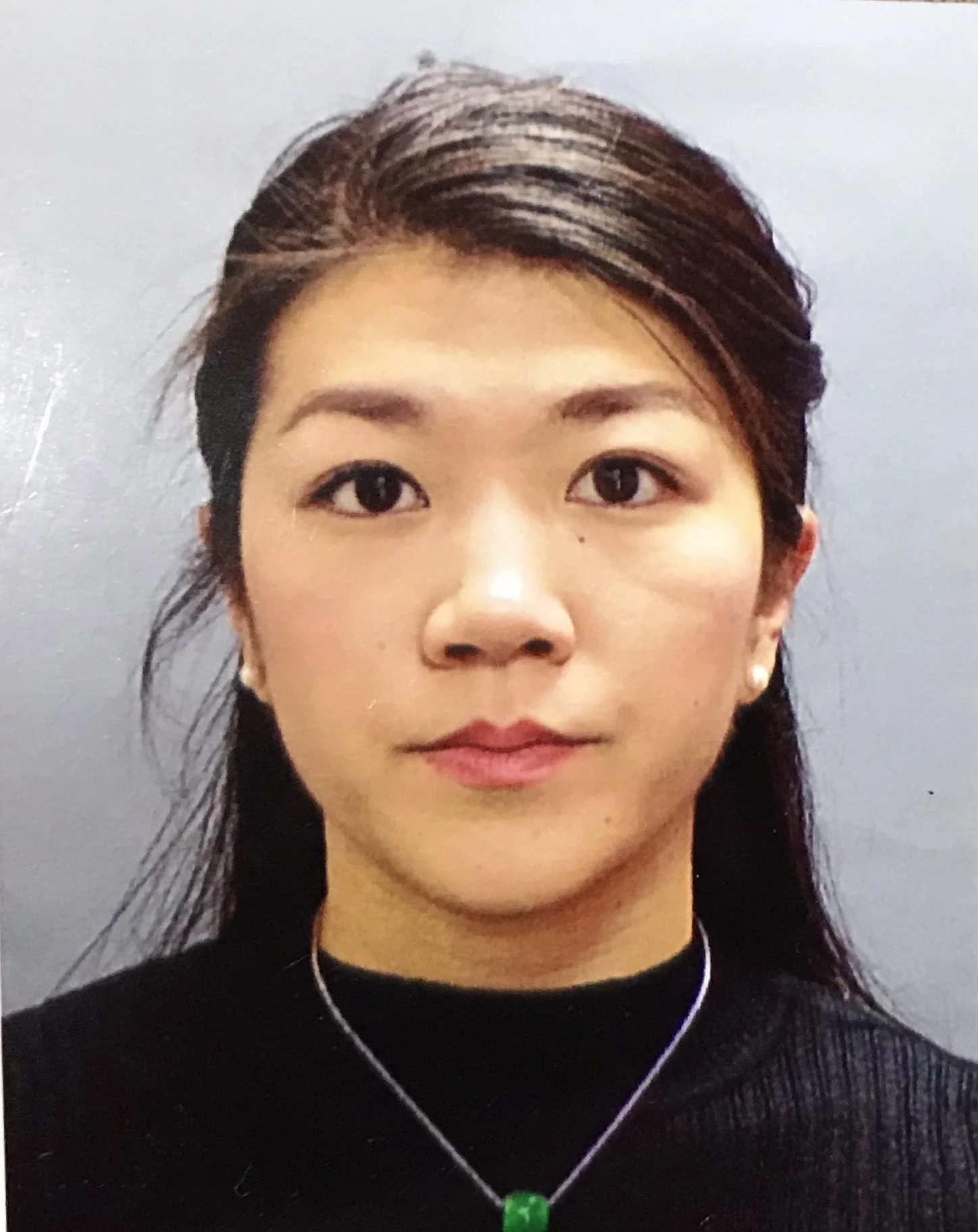 Close-up of an Asian woman with dark hair partially tied back, wearing a black turtleneck and pearl earrings, with earphones and a necklace with a green pendant, standing against a light-colored background.