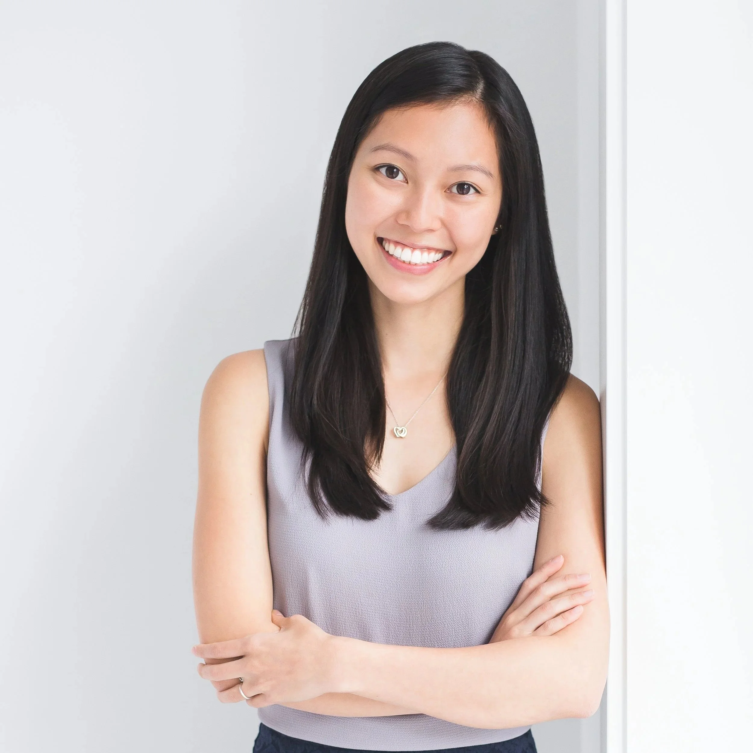 Close-up of a smiling woman with long dark hair, wearing a sleeveless lavender top, standing against a plain white wall.
