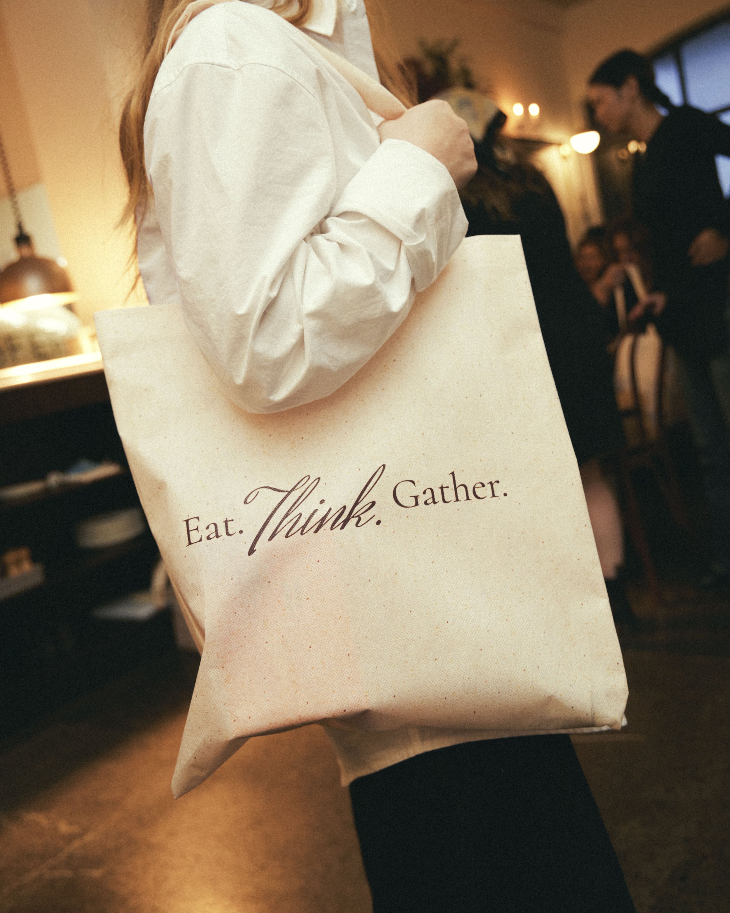A person holding a beige paper bag with the words 'Eat. Think. Gather.' written on it, in a warmly lit indoor setting with other people visible in the background.