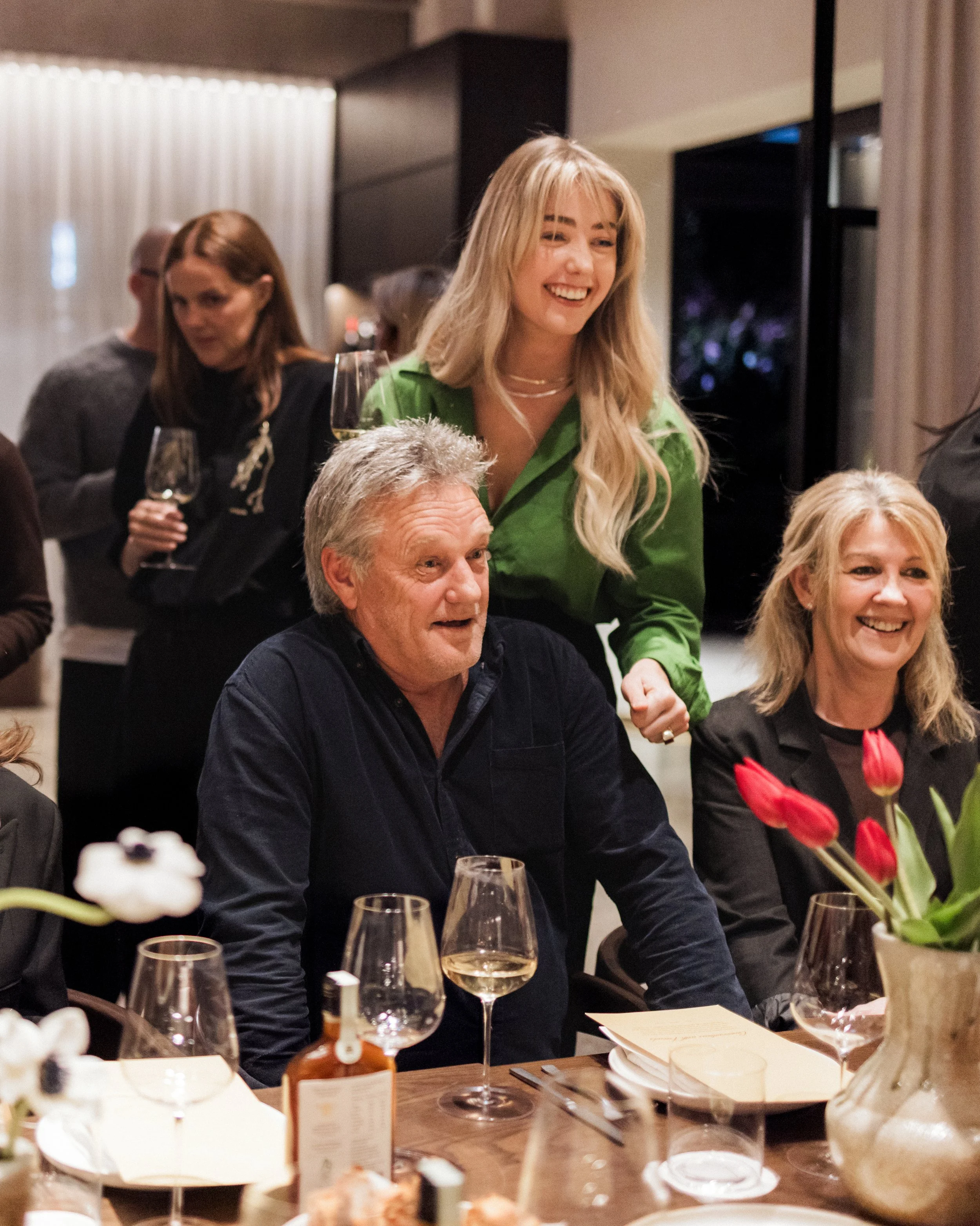 People at dinner party with wine glasses, flowers, and table settings, smiling and socializing in a warmly lit room.