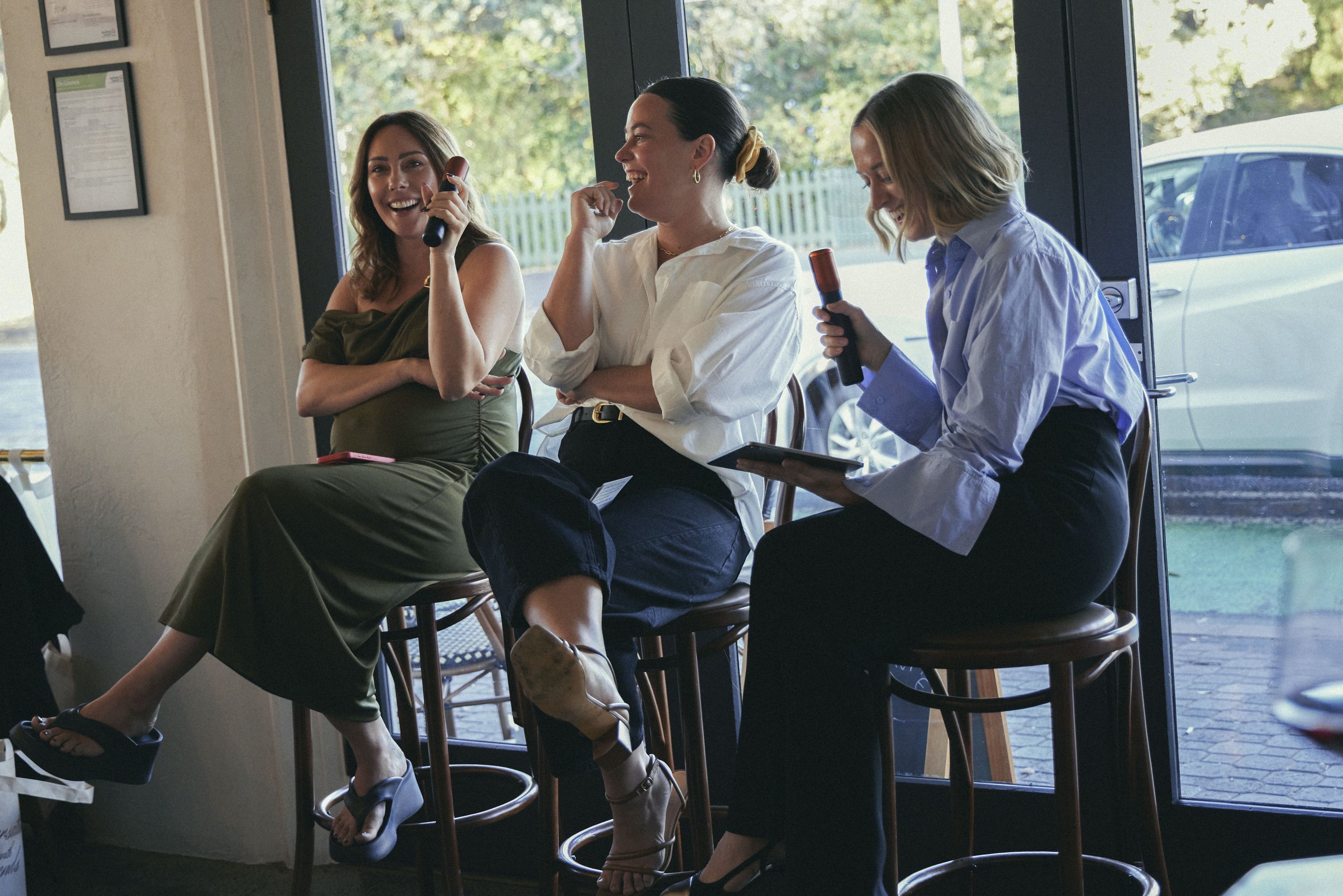 Three women sitting at a high table near a large window, laughing and smiling, with one holding a tablet and the others holding drinks in hand.