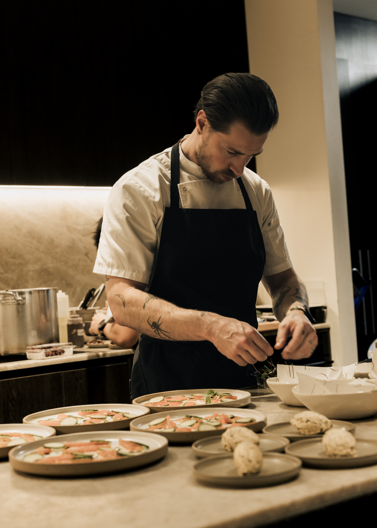 A male chef with tattoos on his arms and slicked-back dark hair preparing dishes in a restaurant kitchen. The counter has multiple plates with appetizers, and the chef is focused on arranging food.