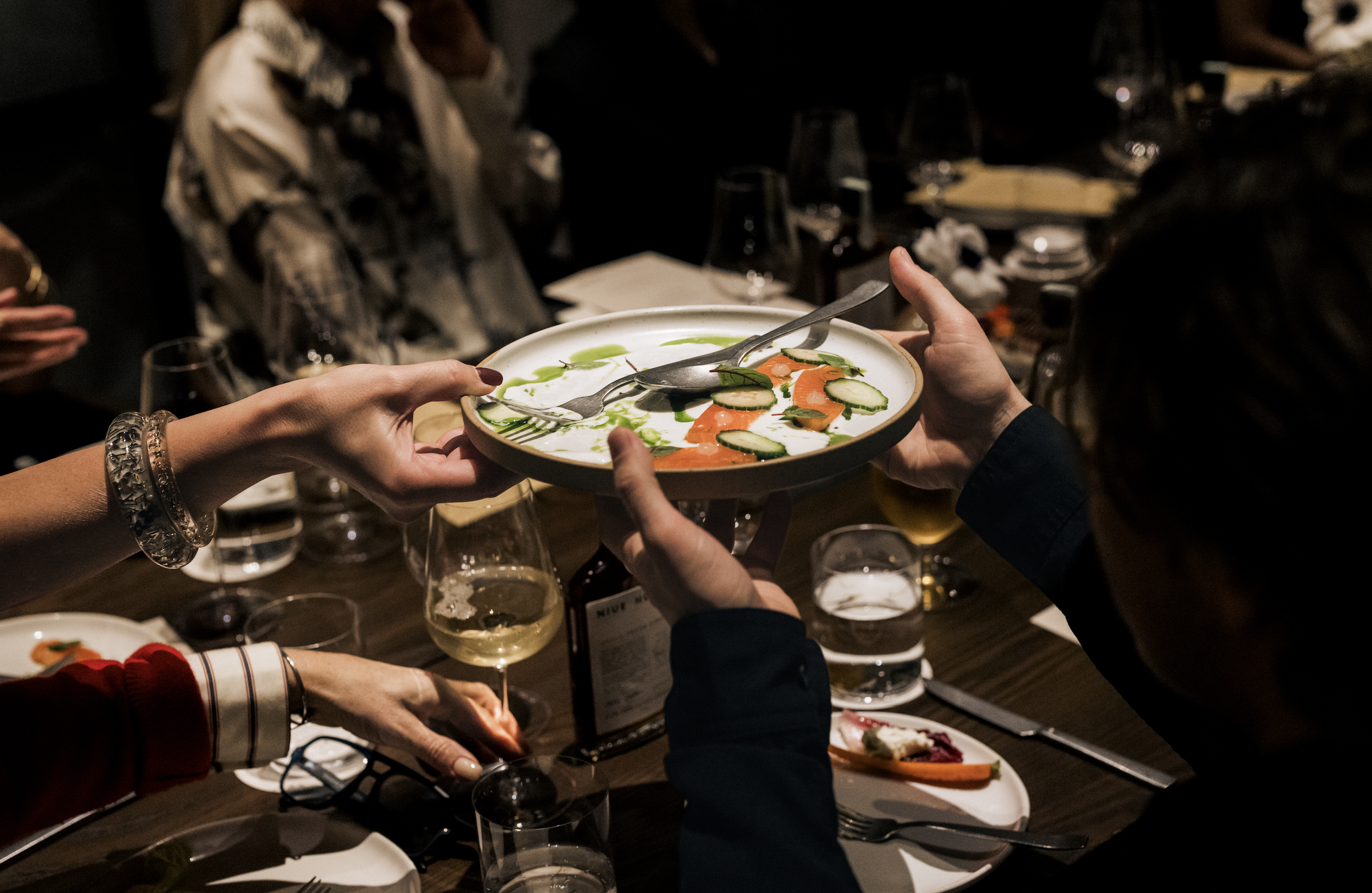 People at a dinner party passing a plate with sliced cucumber and salmon, garnished with green sauce, around a table with wine glasses and plates.
