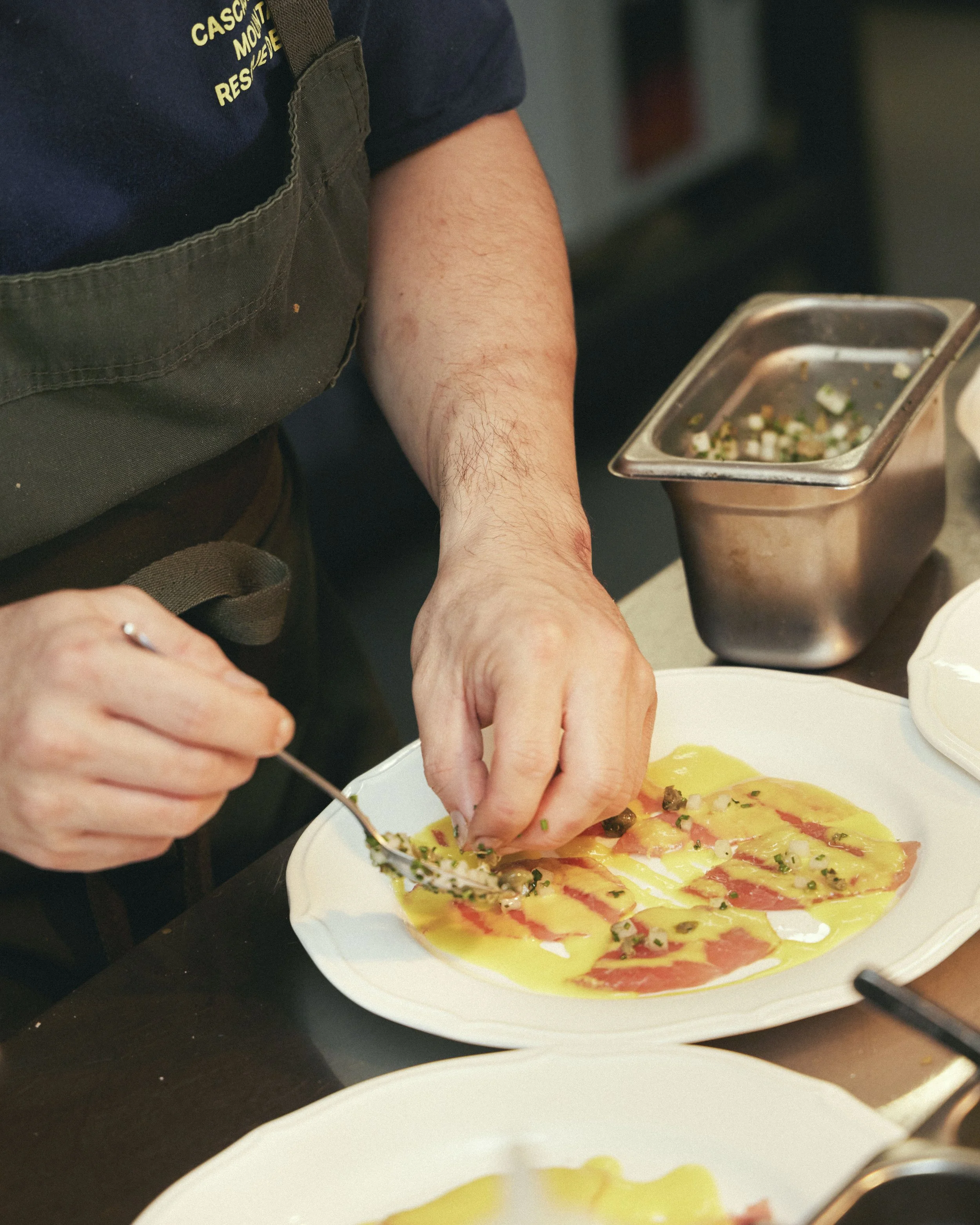 A chef in a dark apron and navy shirt preparing a pizza with toppings on a white plate.