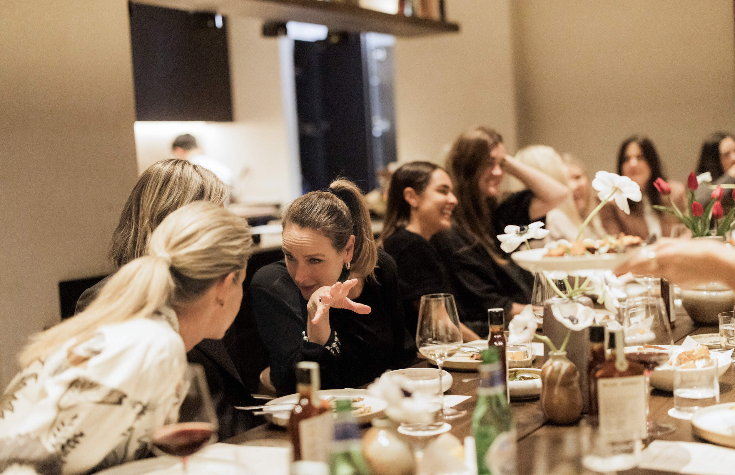 People sitting at a long dinner table, engaged in conversation, with dishes, drinks, and flowers, in an indoor setting.