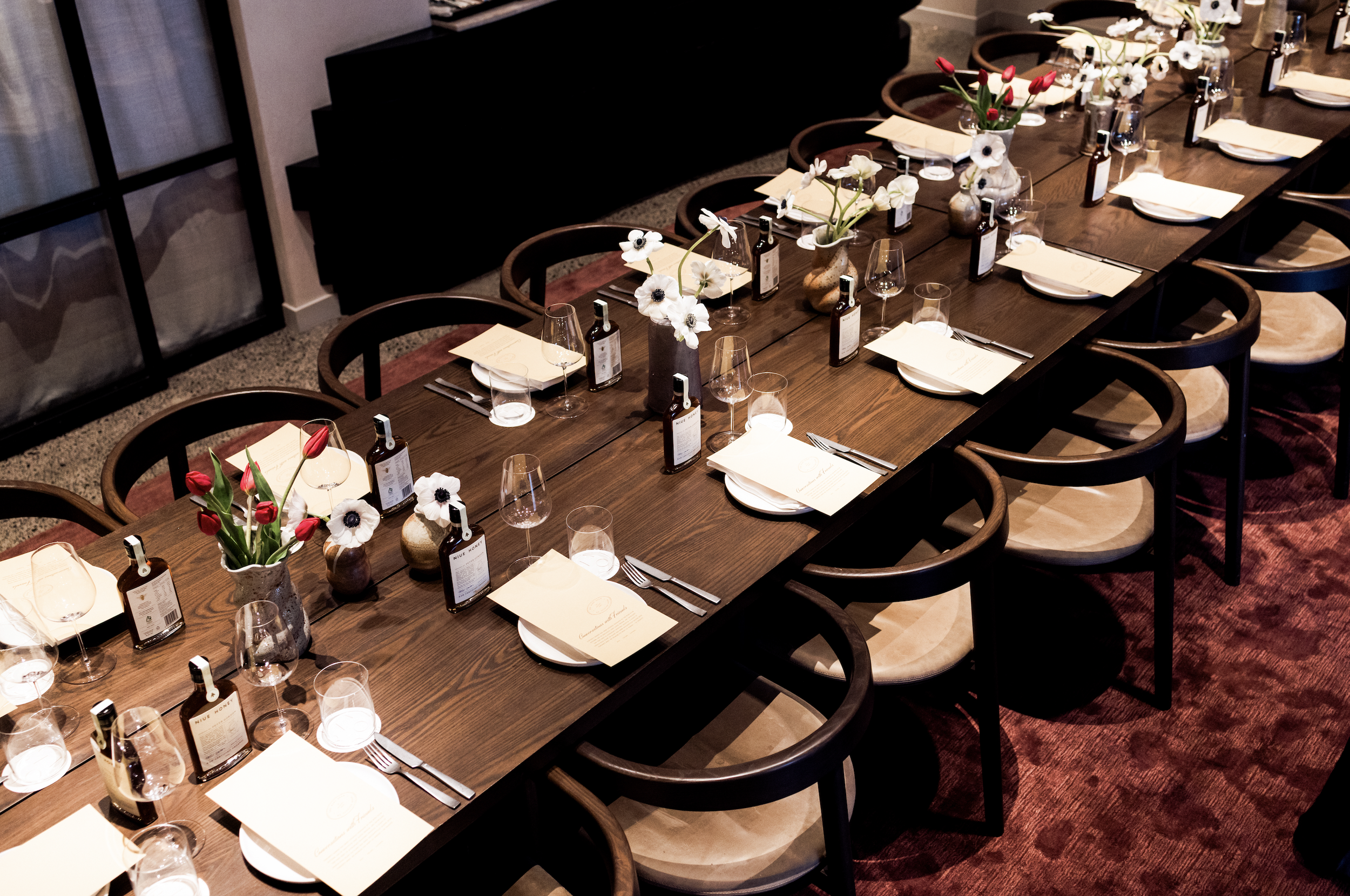 A long dining table set for a meal with individual place settings, including wine glasses, napkins, and silverware. The table has multiple small vases with white and red flowers.