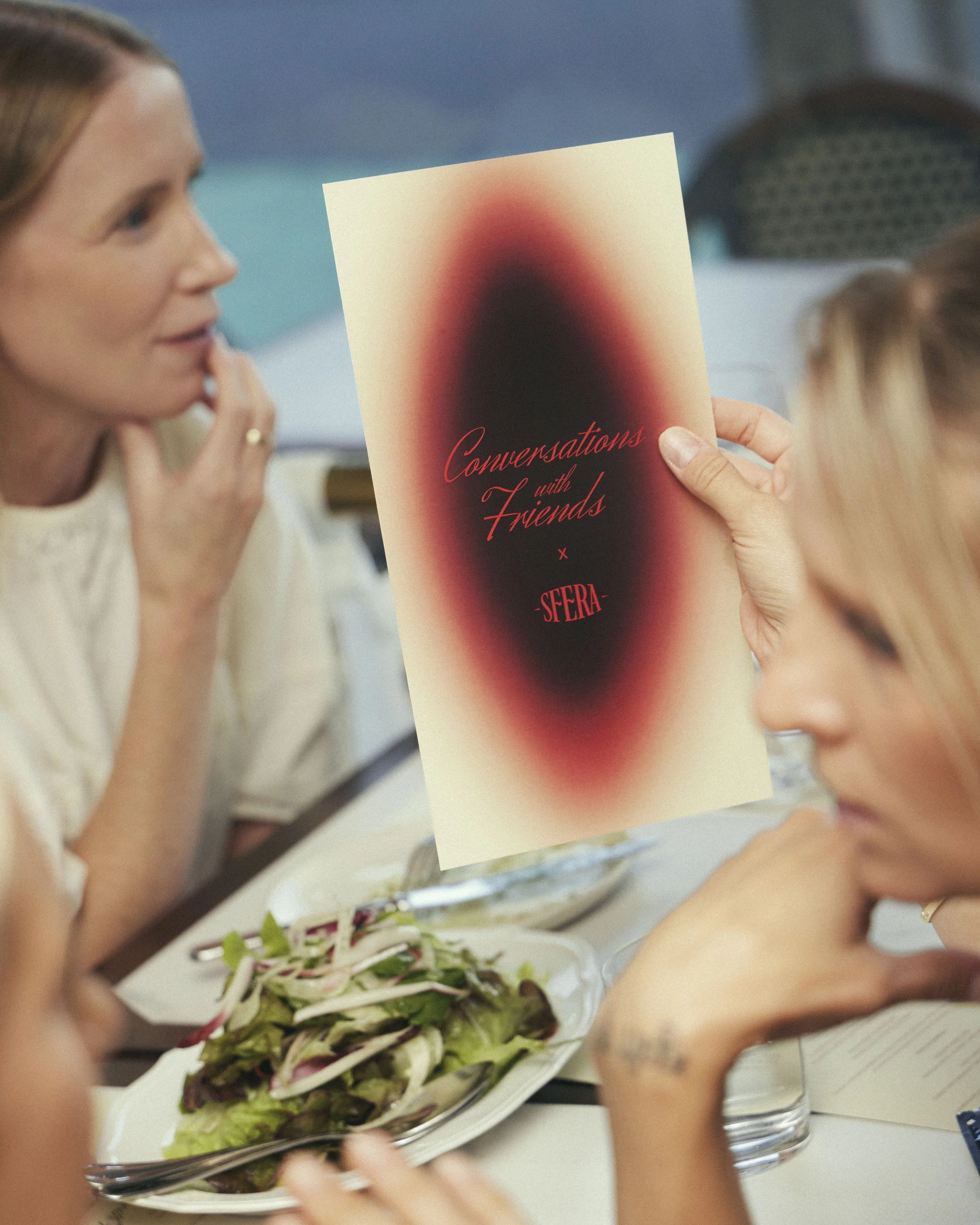 People sitting around a table, one holding a booklet titled 'Conversations with Friends,' with a salad on the table in front of them.