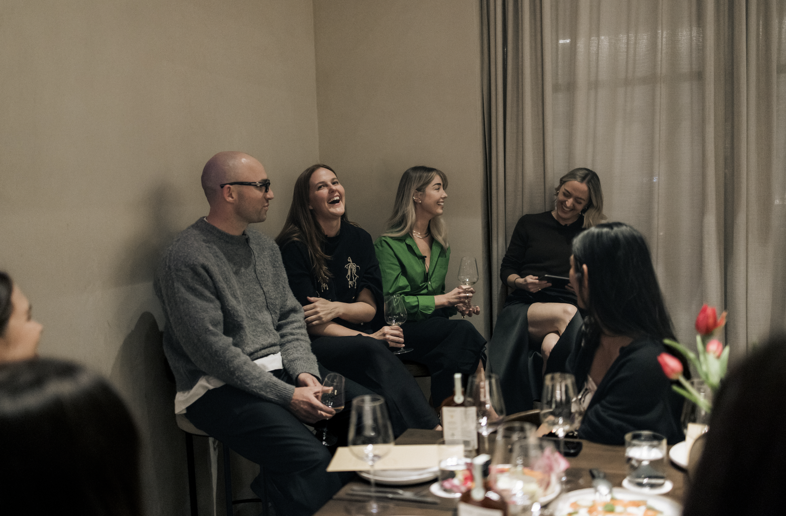 Group of people sitting and laughing during a social gathering or dinner party in a warmly lit room with cream-colored walls and long curtains.