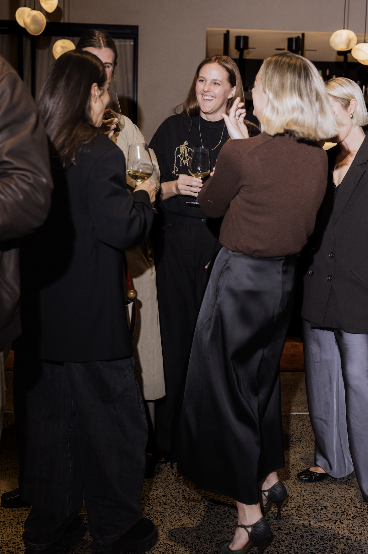 Group of people socializing at a bar or restaurant, holding wine glasses and engaging in conversation, indoors with warm lighting.