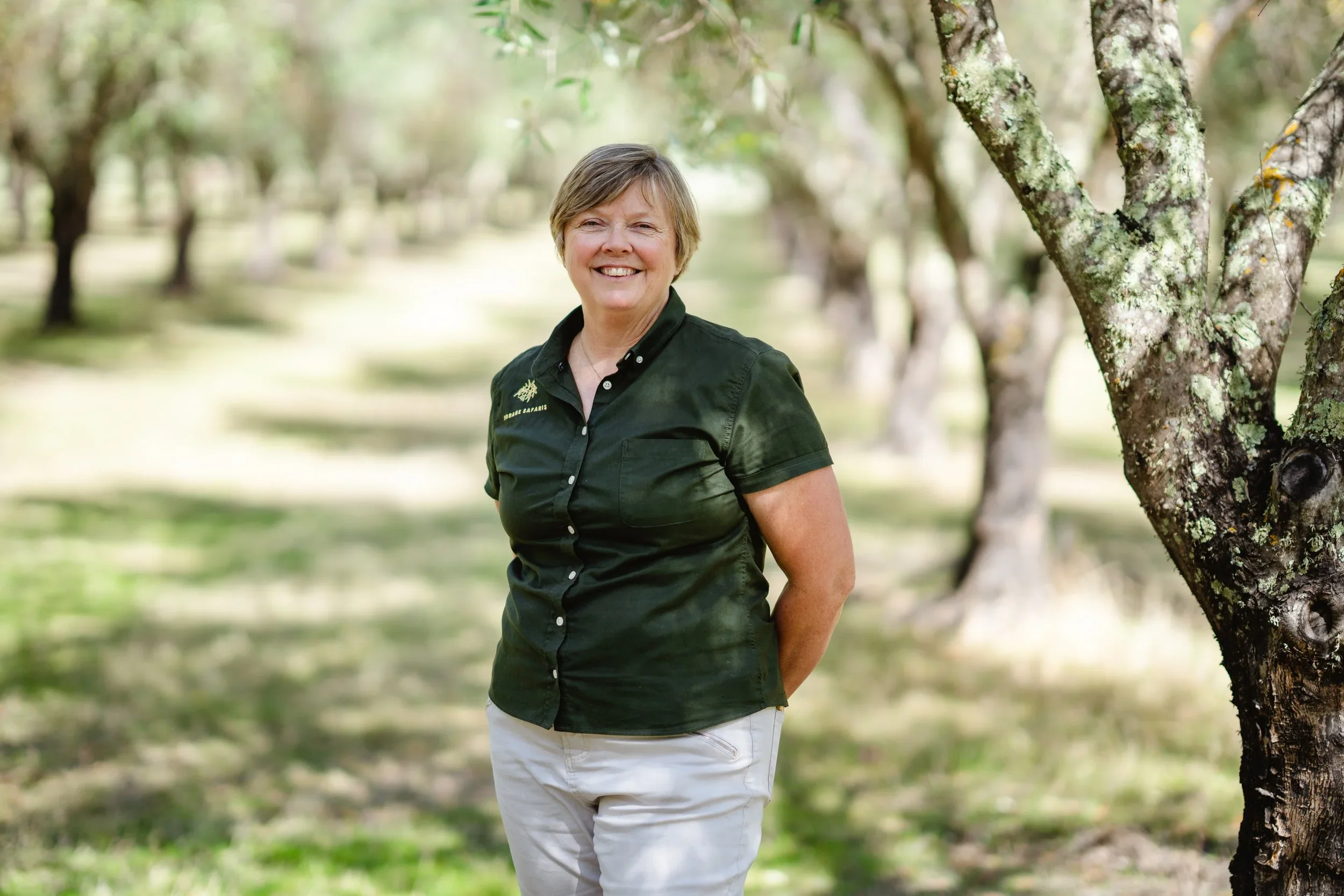 A woman with short hair, wearing a dark green shirt with a logo, smiling outdoors near a tree, in a natural setting with blurred green foliage in the background.