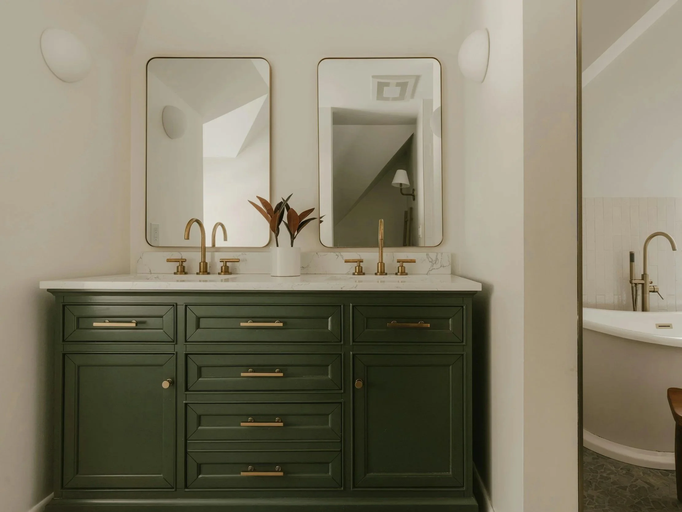 A bathroom vanity with a white countertop, green cabinetry with gold hardware, two oval mirrors, and three brass faucets. There is a plant on the countertop and a partial view of a toilet and shower behind a wall to the right.
