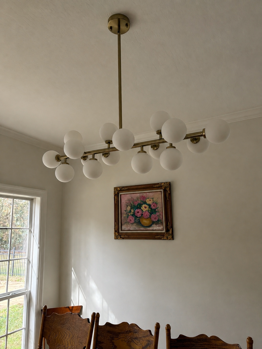 A modern chandelier with multiple white globes hangs from a beige ceiling in a dining room. There is a framed floral painting on the wall and a window letting in natural light.