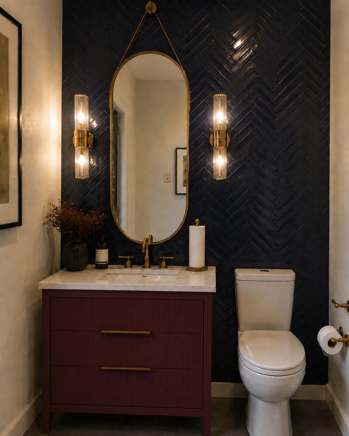 A modern bathroom featuring a dark herringbone tiled wall, an oval mirror with gold frame, gold wall sconces, a white marble countertop with a burgundy vanity, a black vase with dried flowers, a soap dispenser, a paper towel holder, and a white toile