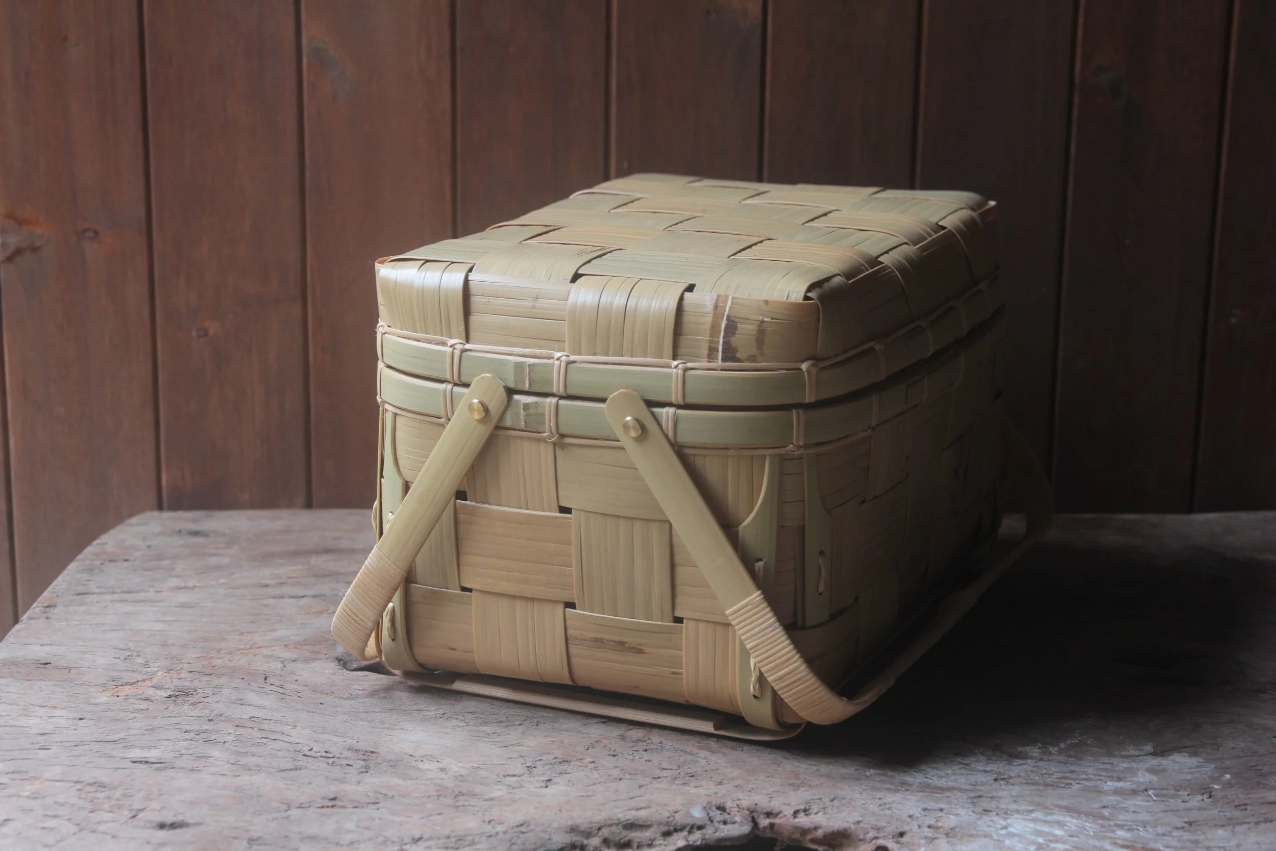 A woven bamboo basket with a lid and a side handle, placed on a rustic wooden table against a dark wooden panel wall.