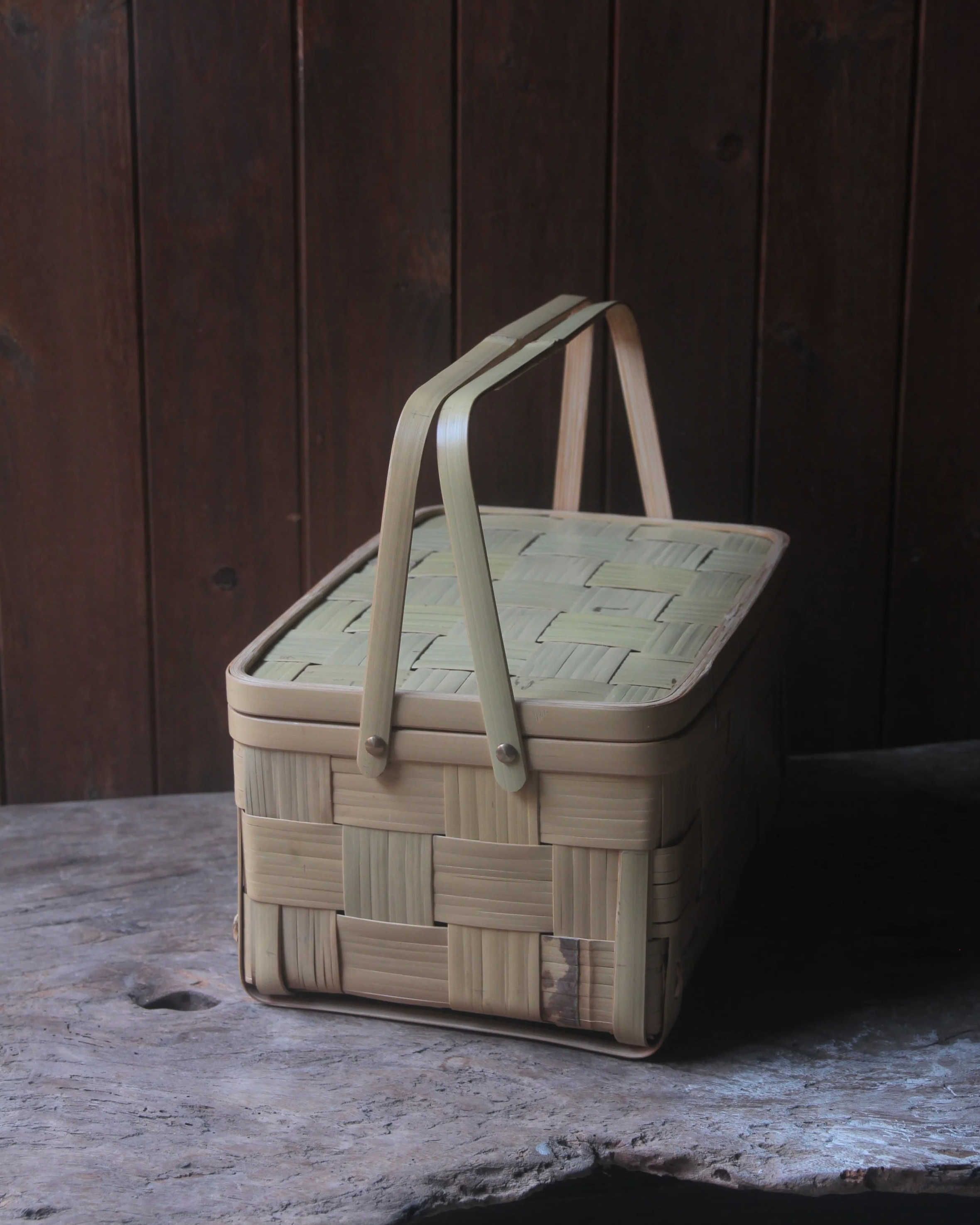 A woven basket with a wooden handle resting on a textured wooden surface against a dark wooden wall background.