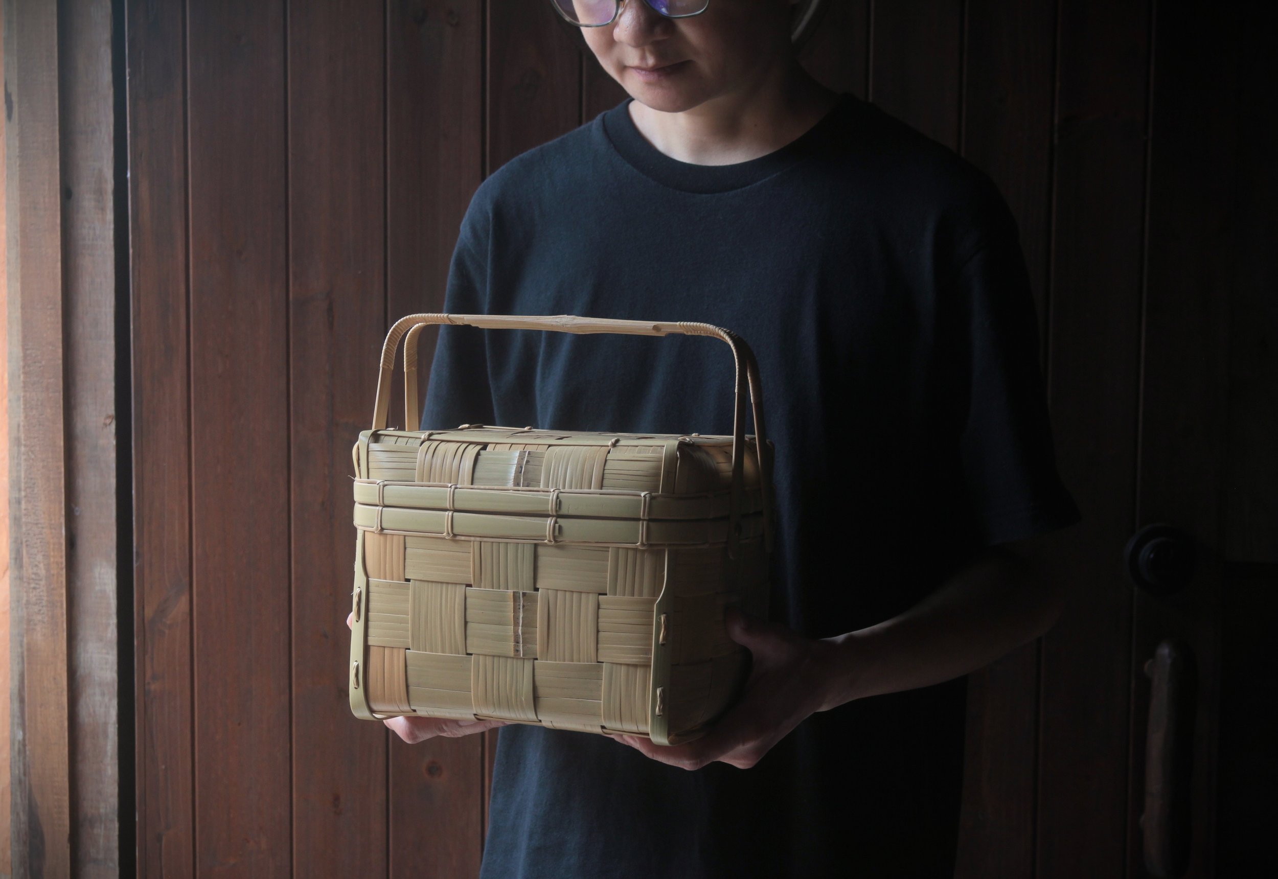 Person wearing glasses and a black shirt holding a woven beige basket, standing against a dark wooden wall.