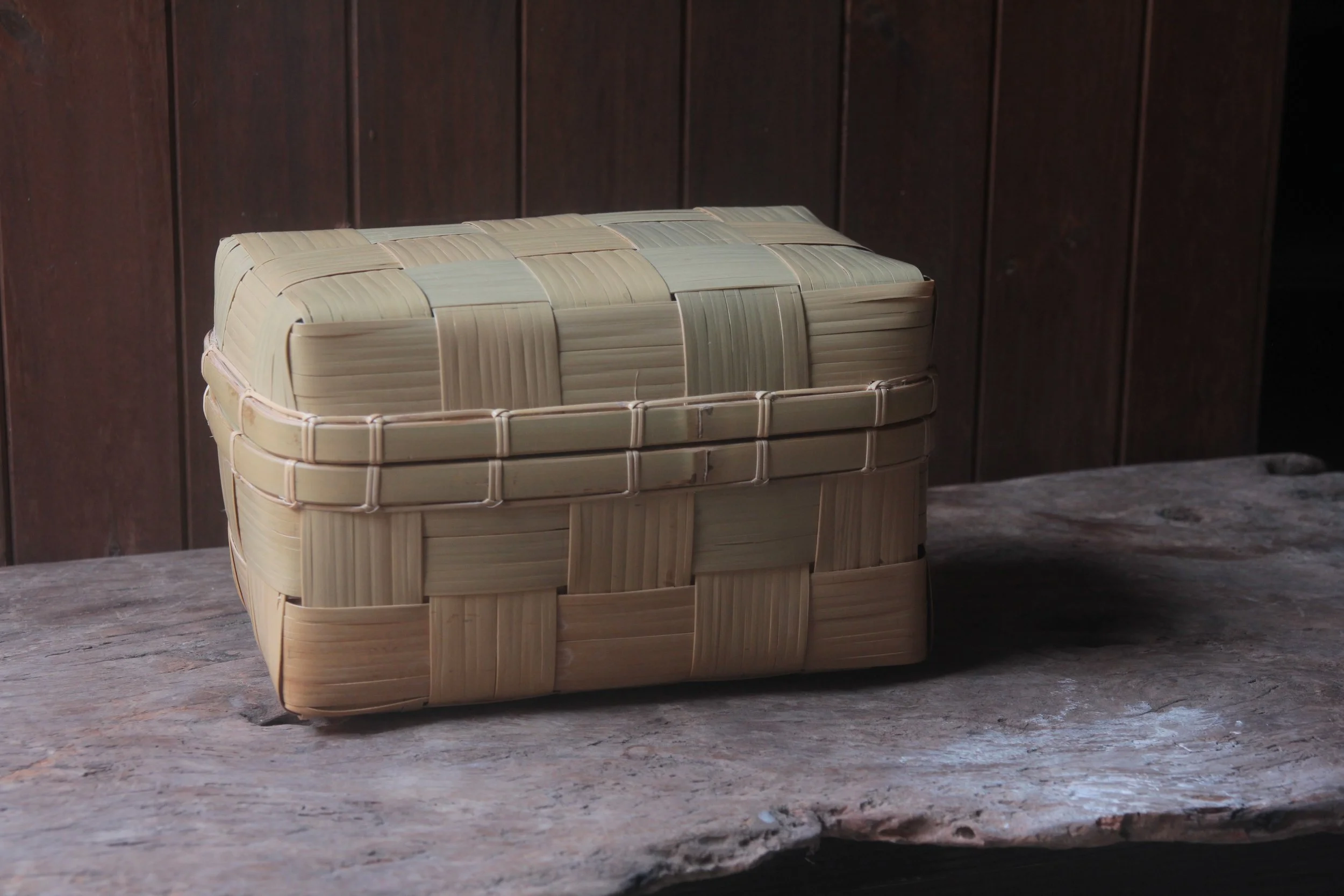 A woven basket made of light-colored strips of material, placed on a wooden surface against a dark wooden paneled wall.