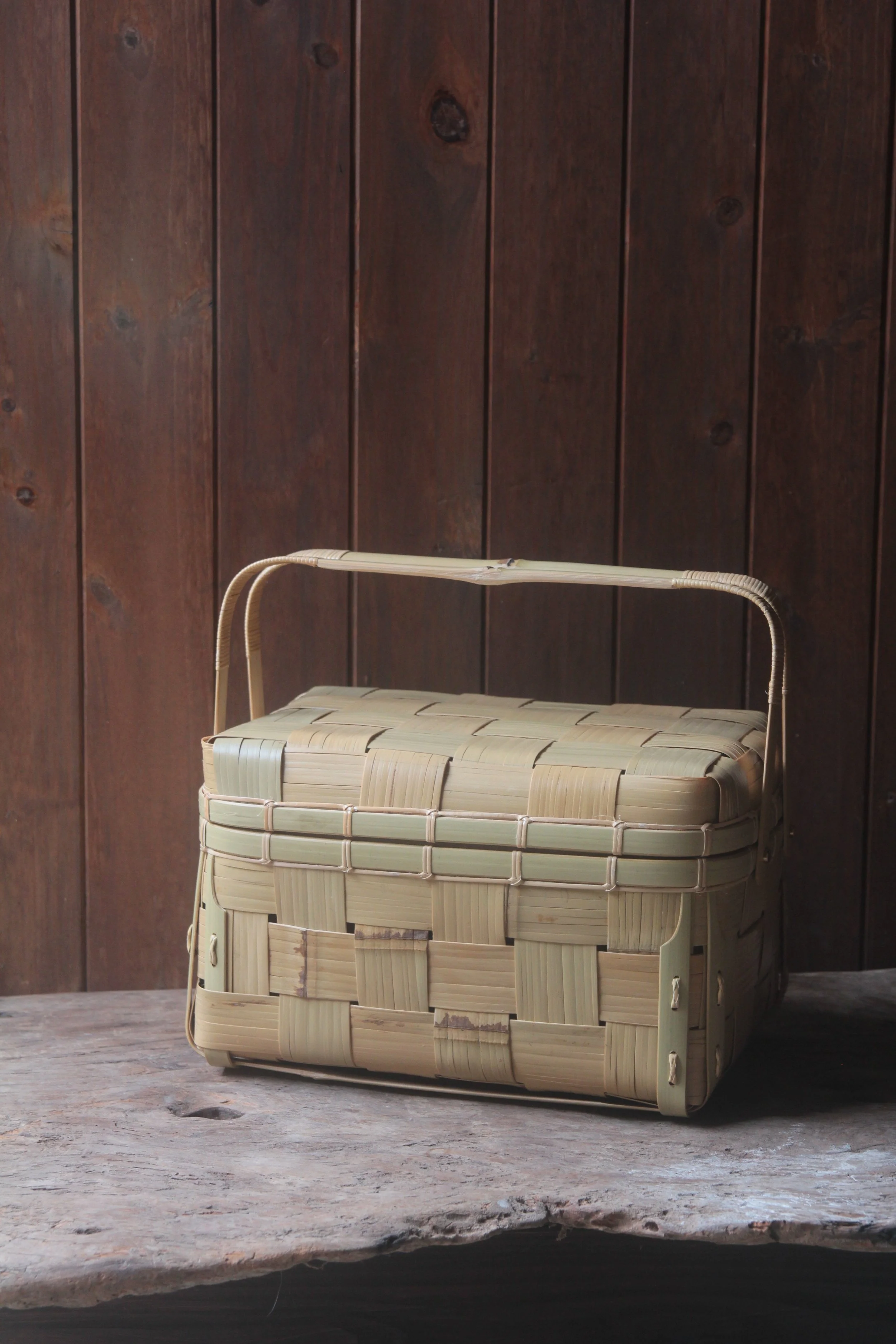 A woven basket with a lid sits on a wooden surface in front of a dark wooden wall.