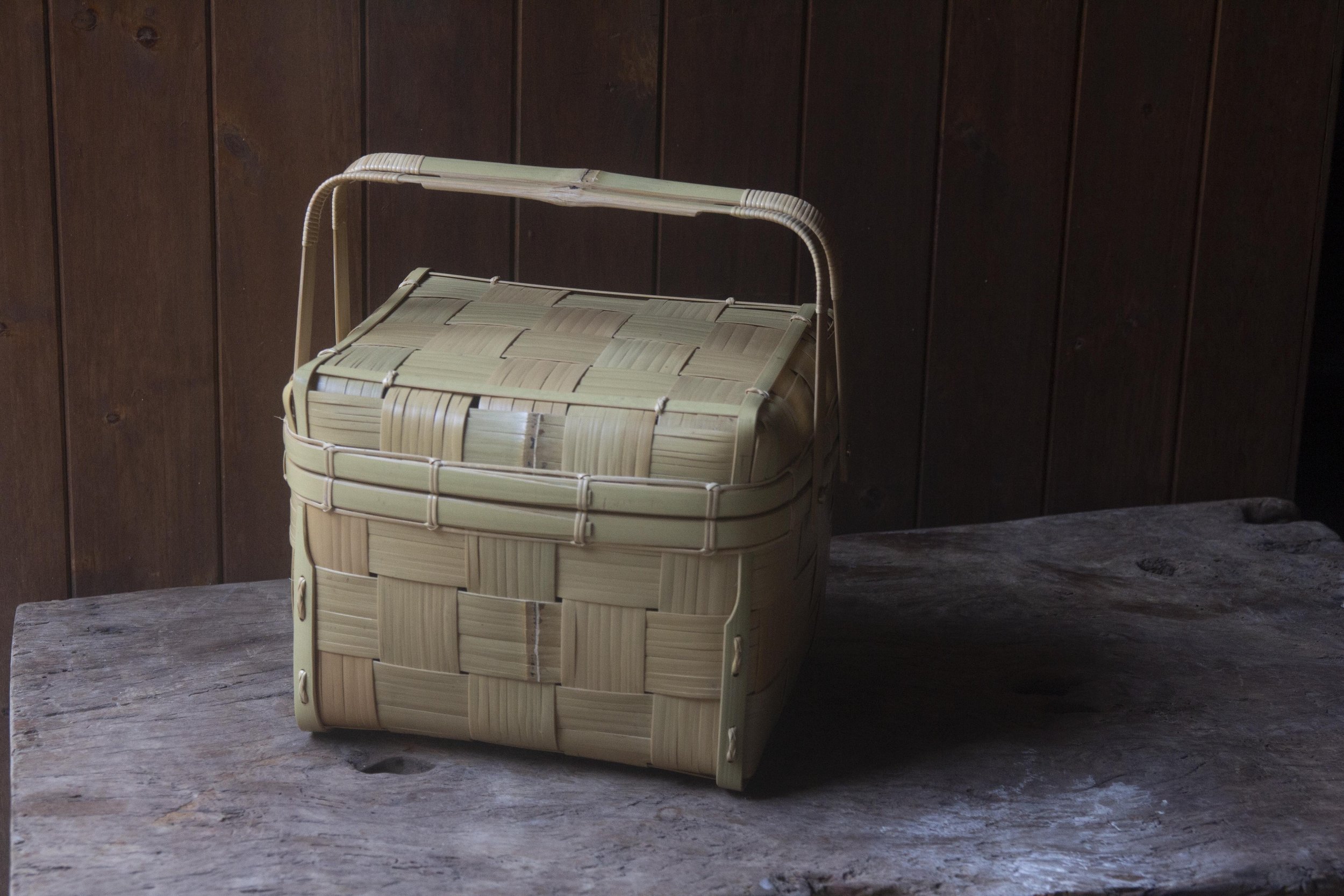 A woven basket with a lid on a wooden surface against a dark wood-paneled wall.