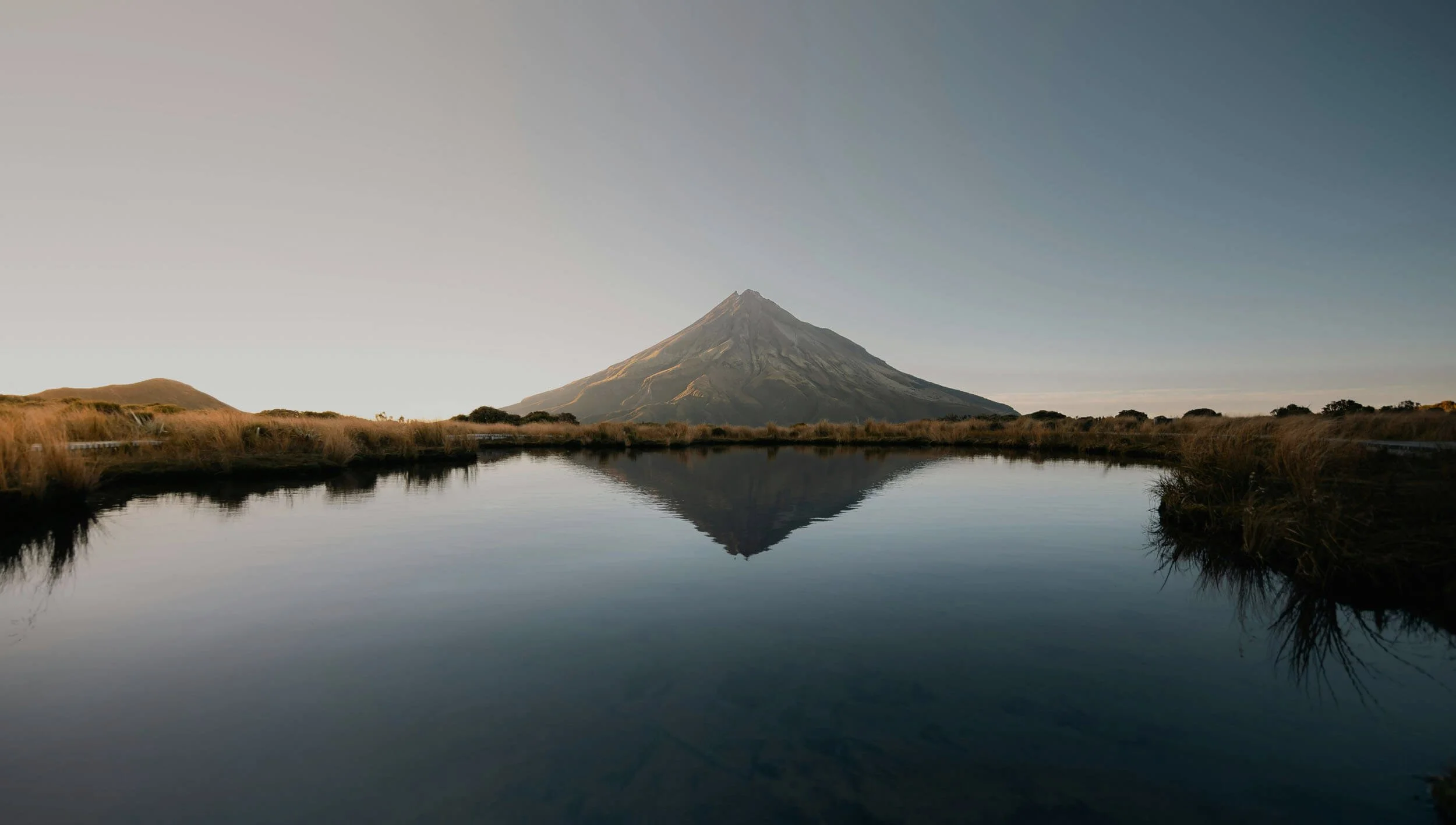 A tall volcano mountain reflected in a calm body of water during early morning or late afternoon with a clear sky.