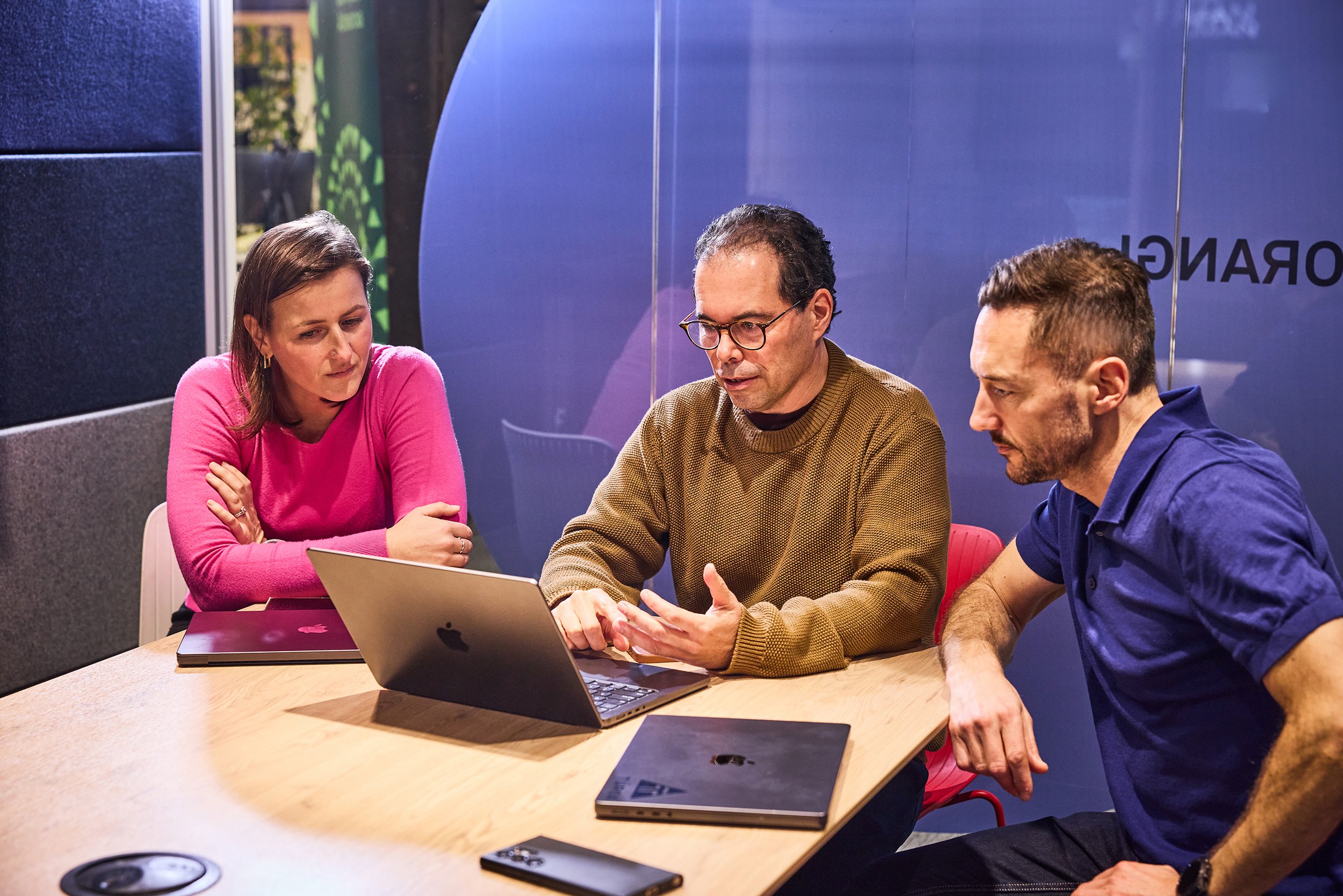 Two people engaging in a conversation at a meeting table in a modern office, with others walking in the background.