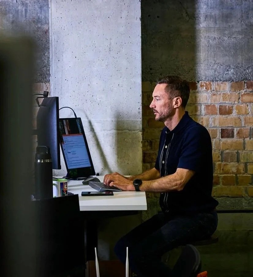 A man working at a desk on a computer in an industrial-style room with exposed brick and concrete walls.