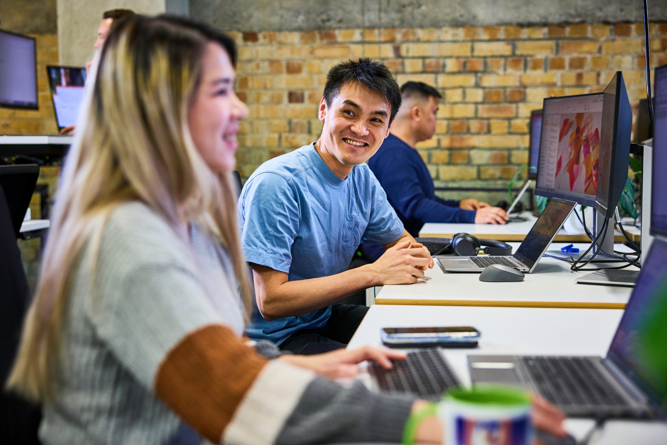 People working at computers in a modern office with exposed brick walls, including a smiling man looking at a woman while working on a laptop.