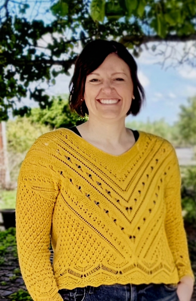A woman with short brown hair smiling outdoors, wearing a yellow crochet sweater, standing in front of a tree with green leaves and a blue sky.