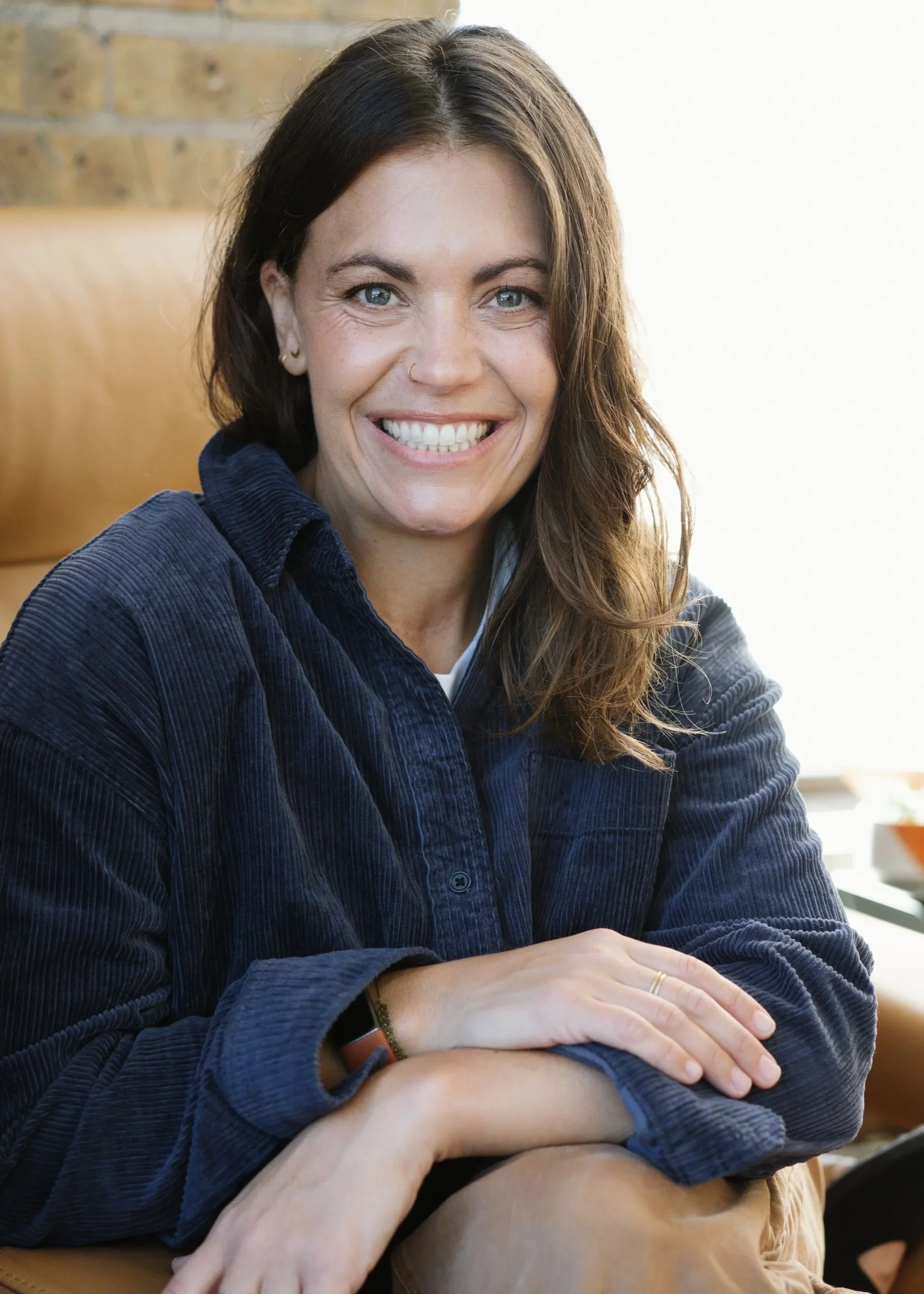A woman with blue eyes, long dark hair, and gold earrings is smiling, sitting indoors near a window with sunlight.