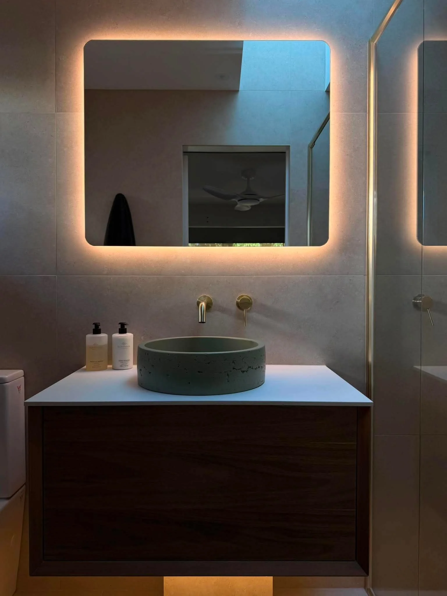Modern bathroom with backlit mirror, green vessel sink, wooden vanity, two soap dispensers, and a white countertop.