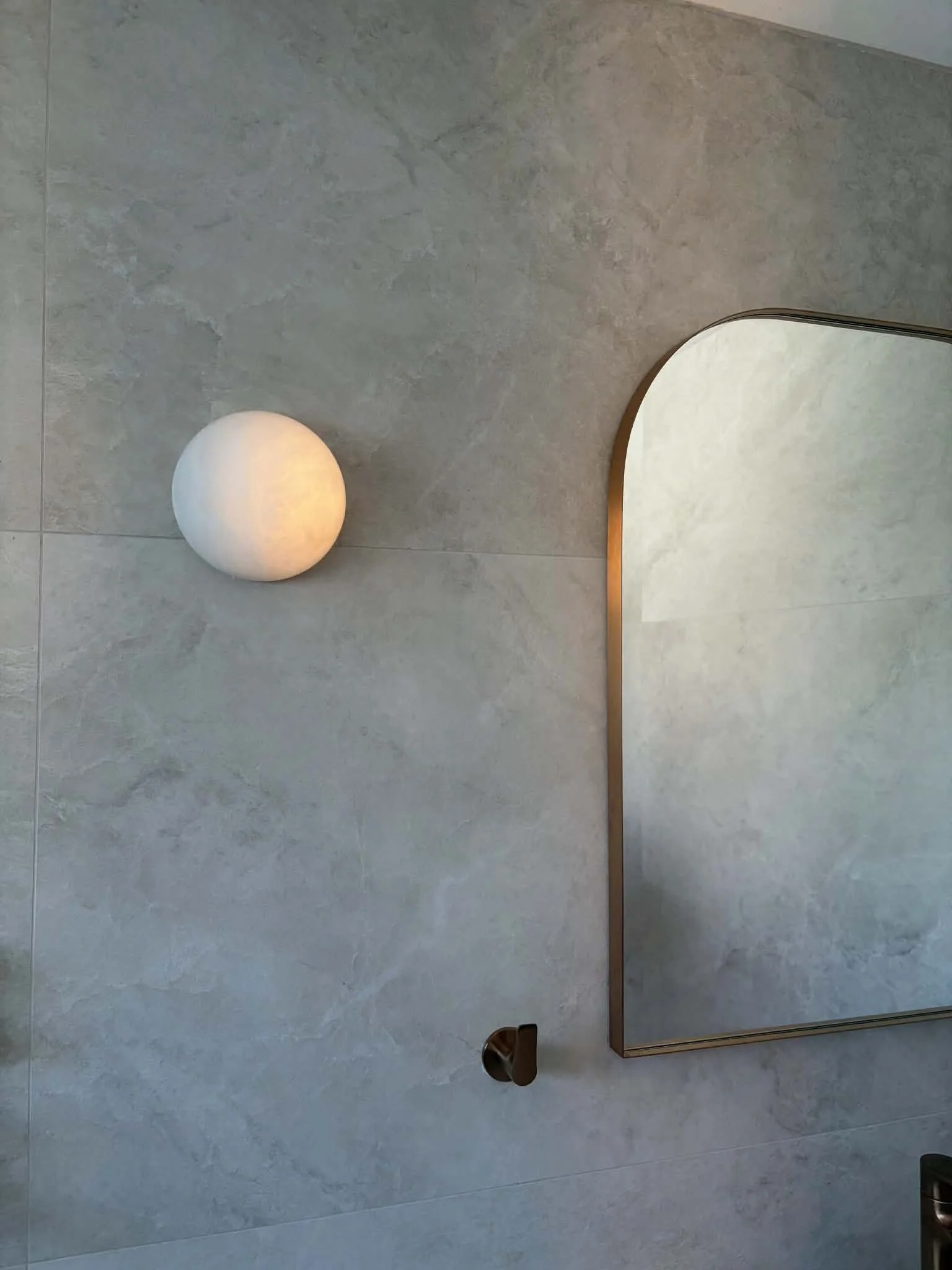 Interior of a modern bathroom with a round wall-mounted light fixture and a mirror with a gold-colored frame reflecting a gray textured wall.